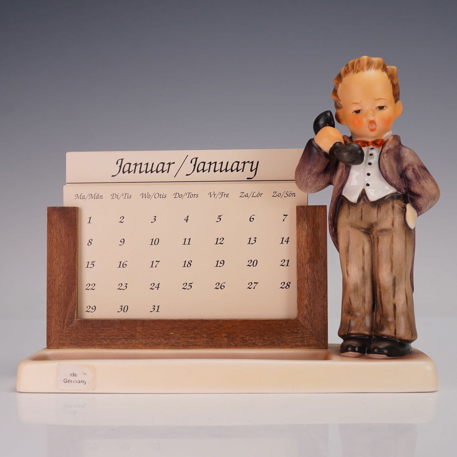 Goebel Hummel Perpetual Calendar, Hello Chef (HUM788/A): A charming porcelain figurine depicting a young boy dressed as a butler holding a telephone, accompanied by a wooden slot designed to hold perpetual calendar cards. The calendar includes interchangeab