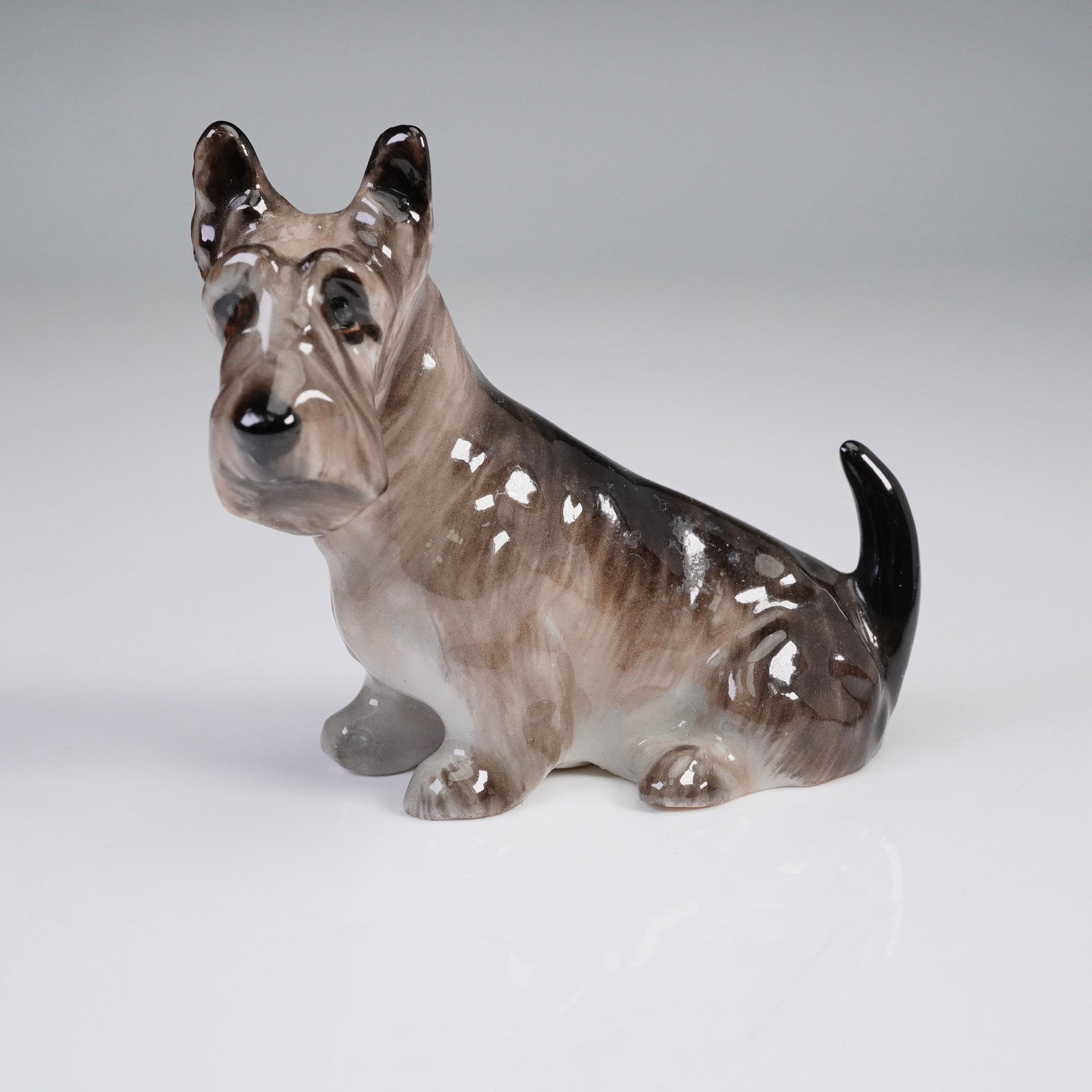 Royal Doulton Animal Figurine, Seated Scottish Terrier, K18: A finely crafted porcelain figurine by Royal Doulton depicting a seated Scottish Terrier. The dog is rendered with lifelike detail in shaded tones of brown, grey, and black with a glossy finish. Royal