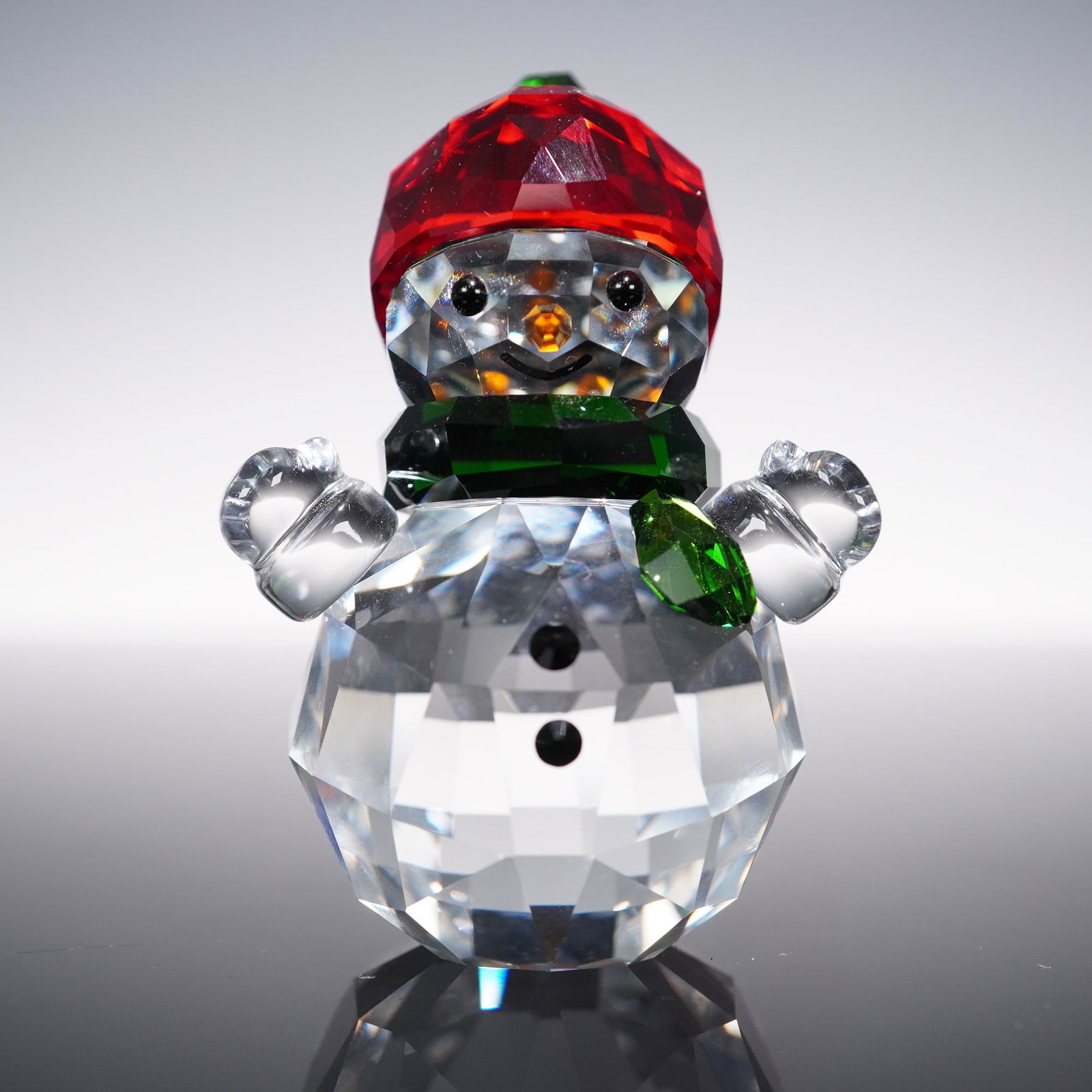 Swarovski Crystal Figurine, Snowman With Red Hat 5288205: A charming faceted crystal snowman adorned with a bright red crystal hat, green crystal scarf, and clear crystal body. The figure features black crystal eyes, buttons, and a cheerful smile, complement