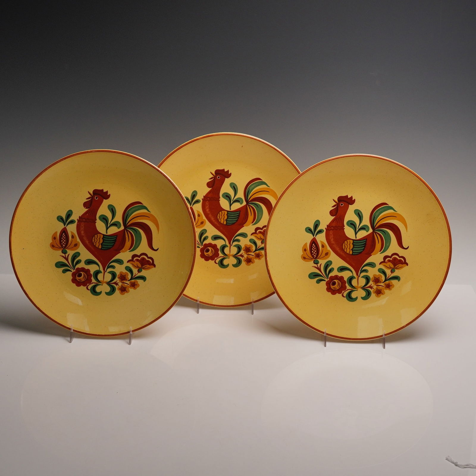 3pc Taylor Smith and Taylor Rooster Dinner Plates: Set of three cheerful golden yellow plates each decorated with a proud rooster in bold shades of red green and orange surrounded by stylized floral accents. The design is lively and folk inspired with