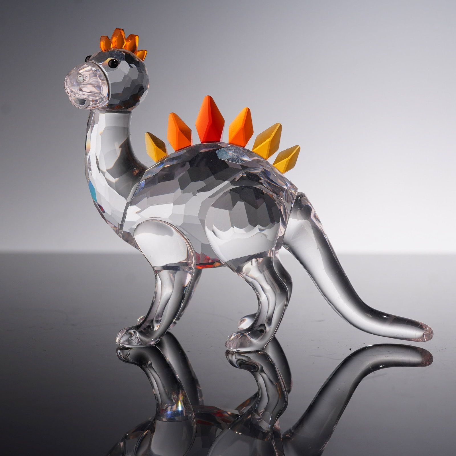 Swarovski Crystal Figurine Dino Dinosaur 268204: This Swarovski crystal figurine titled Dino is part of the Fairy Tales collection. The dinosaur is crafted from clear faceted crystal with vibrant crystal plates and comb ranging in color from yellow 