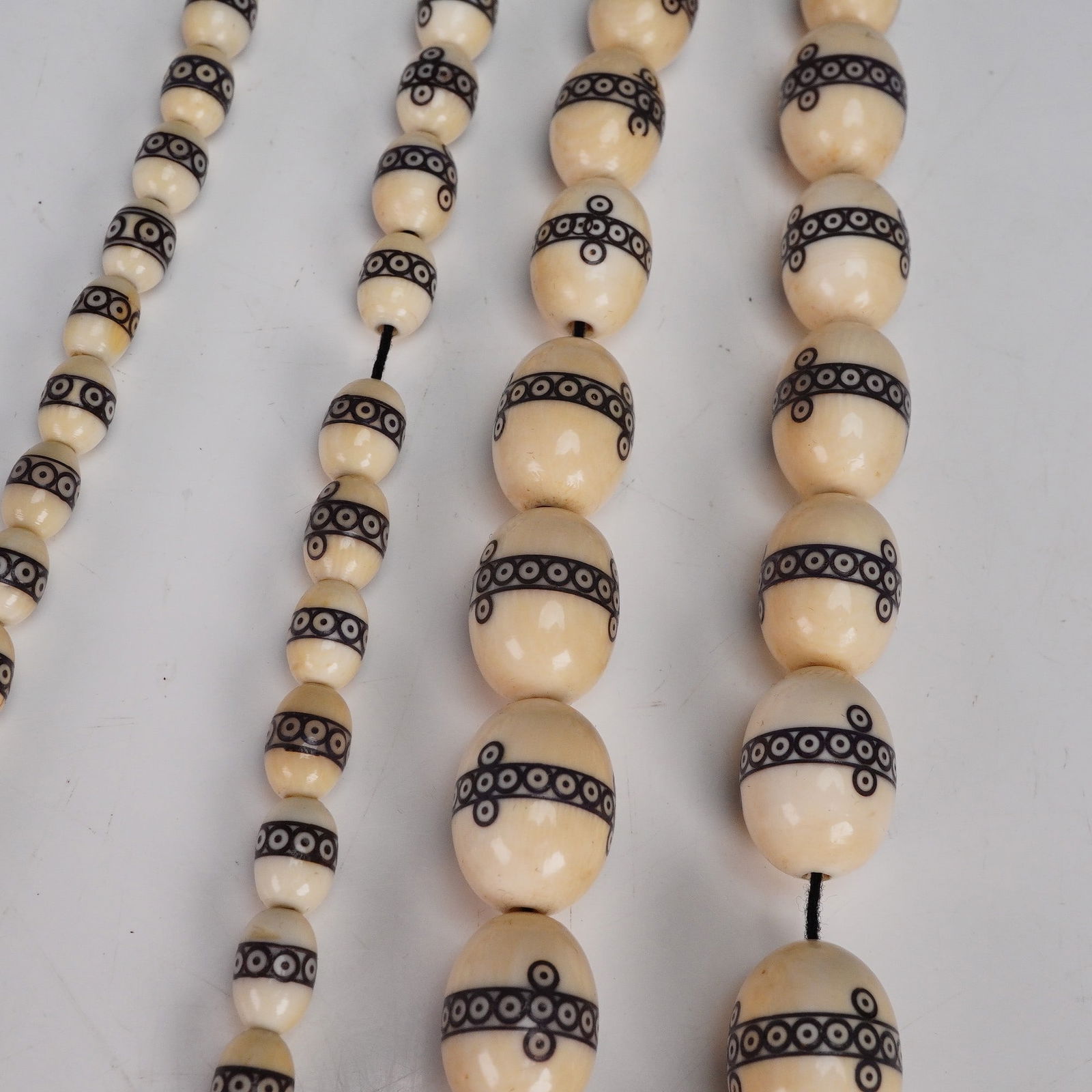 Carved Bone Bead Necklace with Black Inlaid Motifs: A long necklace composed of polished cream-colored carved bone beads, each decorated with a black inlaid band of circle-and-dot motifs. The strand is graduated, with larger beads at the center taperin