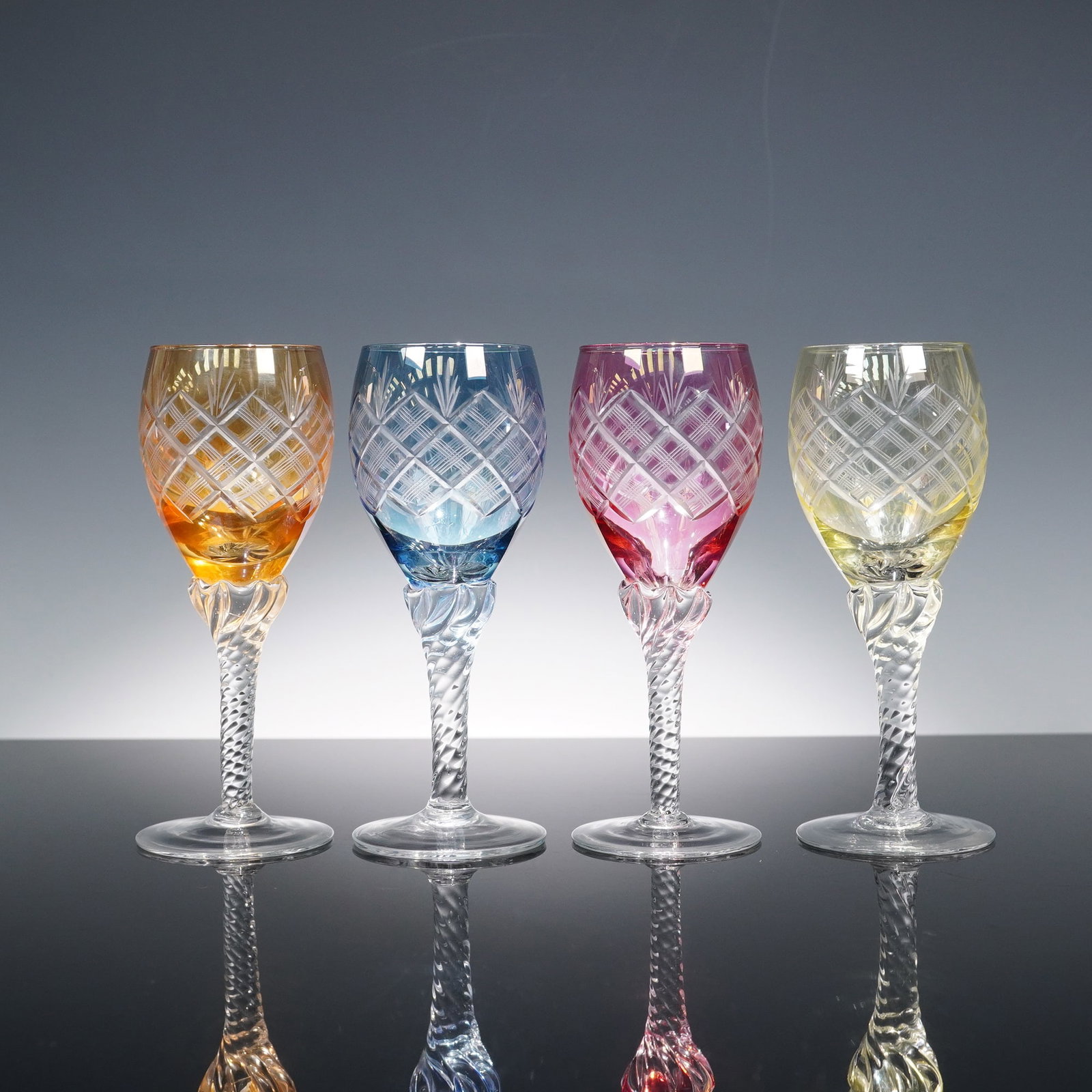 4pc Set of Multicolor Cut to Clear Crystal Wine Glasses: A vibrant set of four cut to clear crystal wine glasses, each in a distinct color: amber, blue, cranberry, and yellow. The bowls feature cross-hatched lattice cuts above faceted detailing, paired with