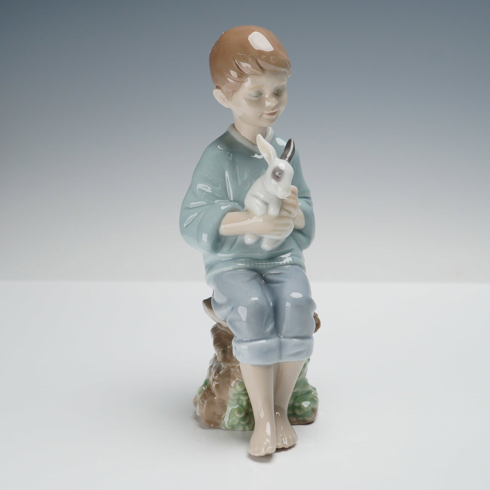 Lladro Porcelain Figurine, Every Bunny Needs Somebody 1008287: A delightful Lladro porcelain figurine depicting a young boy seated on a stack of logs while gently holding a rabbit in his arms. The soft pastel palette of blue, brown, and cream highlights Lladro's 