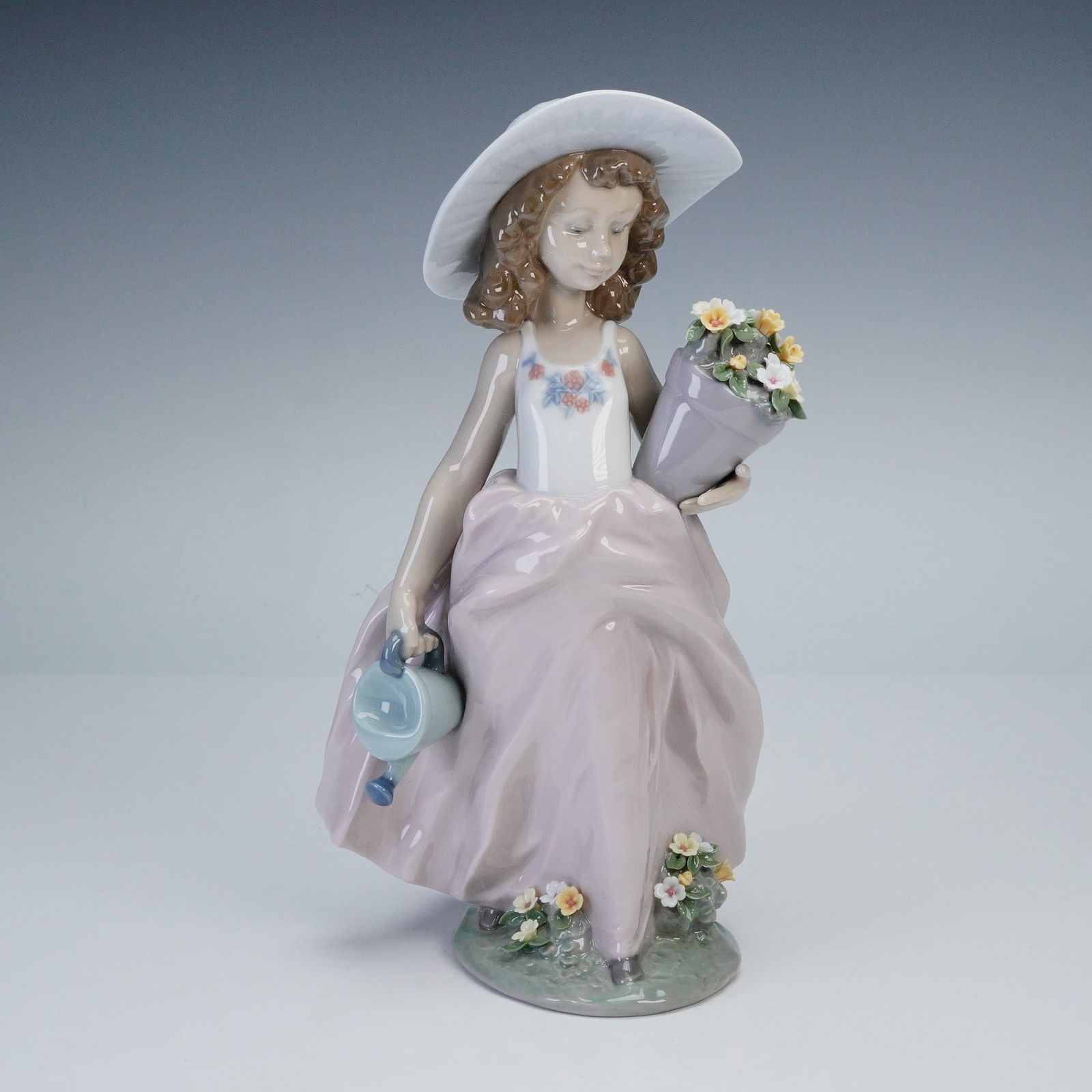 Lladro Society Porcelain Figurine, A Wish Come True 1007676: Features a young girl wearing a wide-brimmed hat, carrying a bouquet of flowers and a watering can, with additional floral details at the base. Finished in soft pastel glazes of lavender, white, and b