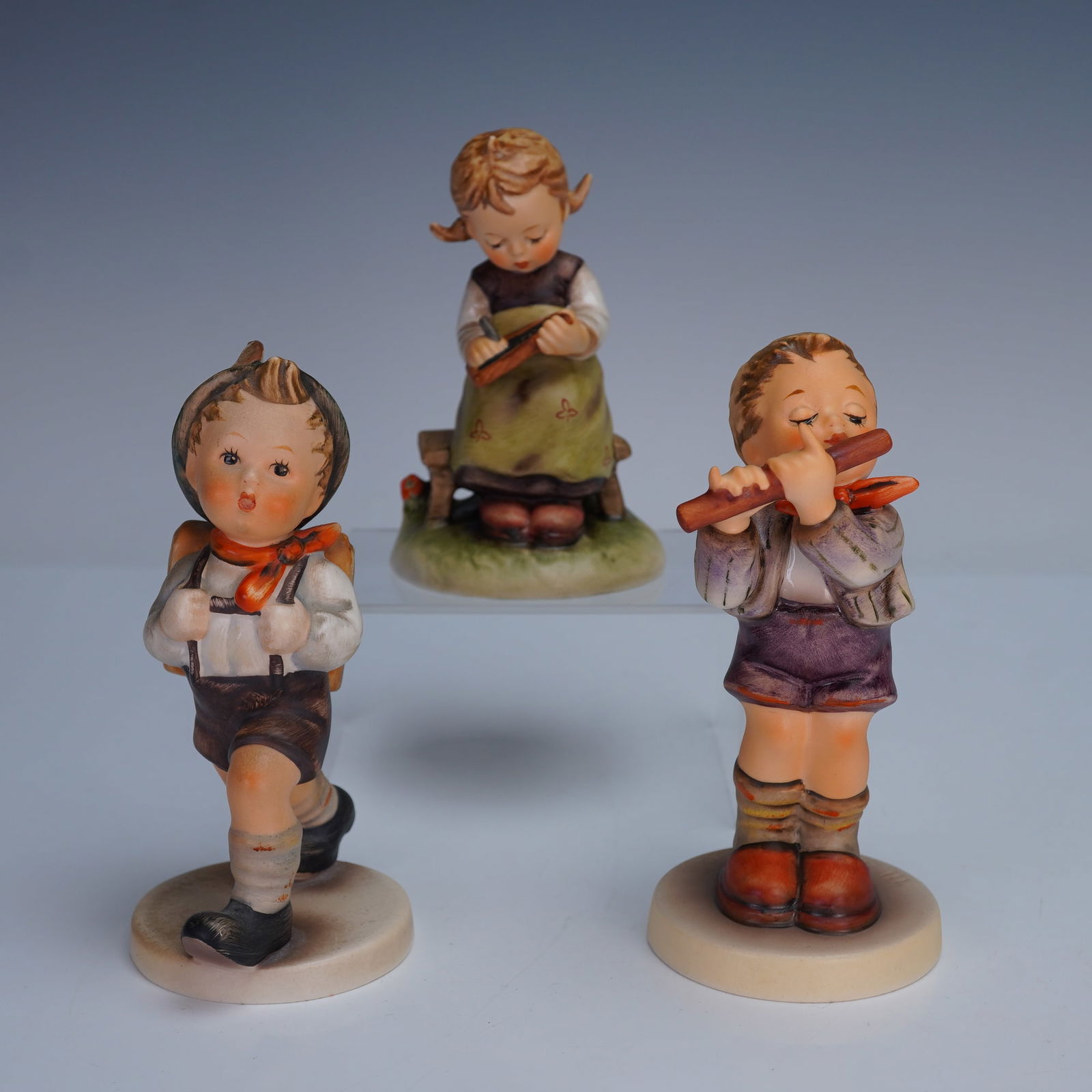 3pc Goebel Hummel Children Figurines, School and Activities: A set of 3 Goebel Hummel porcelain figurines depicting children in everyday activities. One shows a boy walking with his school satchel. Another features a boy playing a flute, base marked "Exclusive