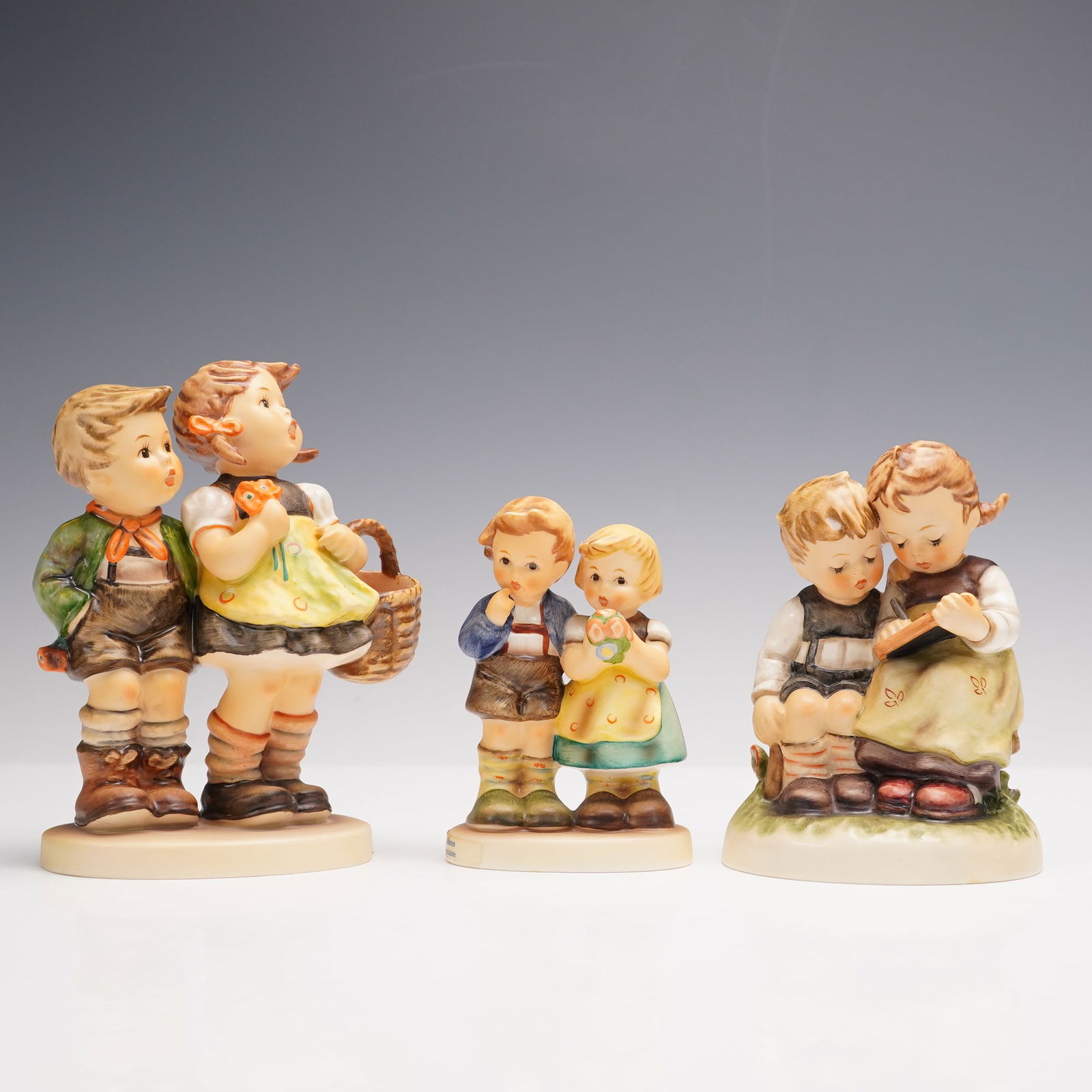 3pc Goebel Hummel Figurines, Children in Pairs: A charming trio of Goebel Hummel porcelain figurines, all hallmarked. The group features a boy and girl walking together with a basket in hand, two youngsters standing side by side with a flower bouqu