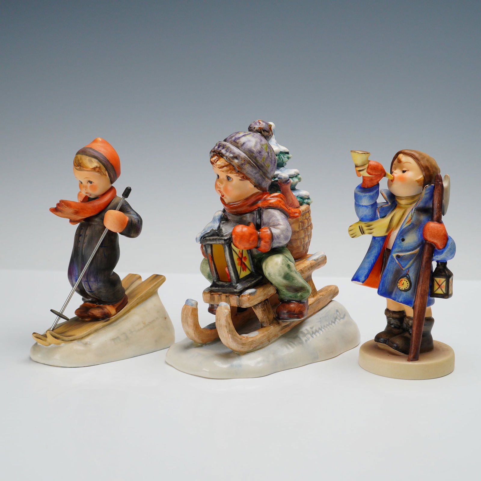 3pc Goebel Hummel Winter Themed Figurines: A delightful winter themed grouping of Goebel Hummel figurines. Skier depicts a young child in a dark snowsuit and bright red scarf, balancing carefully on skis. Hear Ye Hear Ye shows a boy in a blue