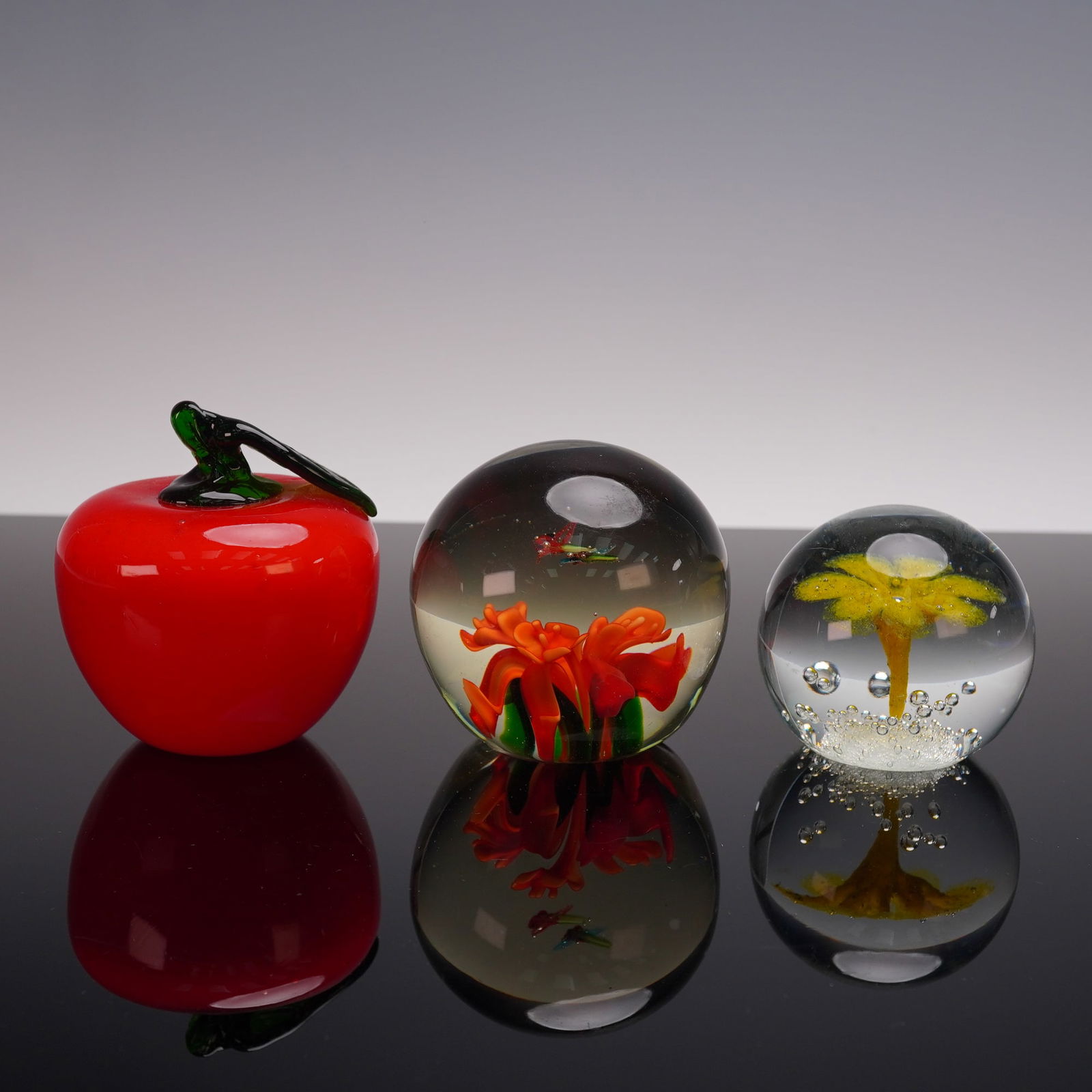 Group of 3 Decorative Art Glass Paperweights- Apple + Red & Yellow Flowers: A charming group of three decorative art glass paperweights, each showcasing vibrant designs. The collection includes a glossy red apple with green stem, a spherical paperweight encasing vivid red flo