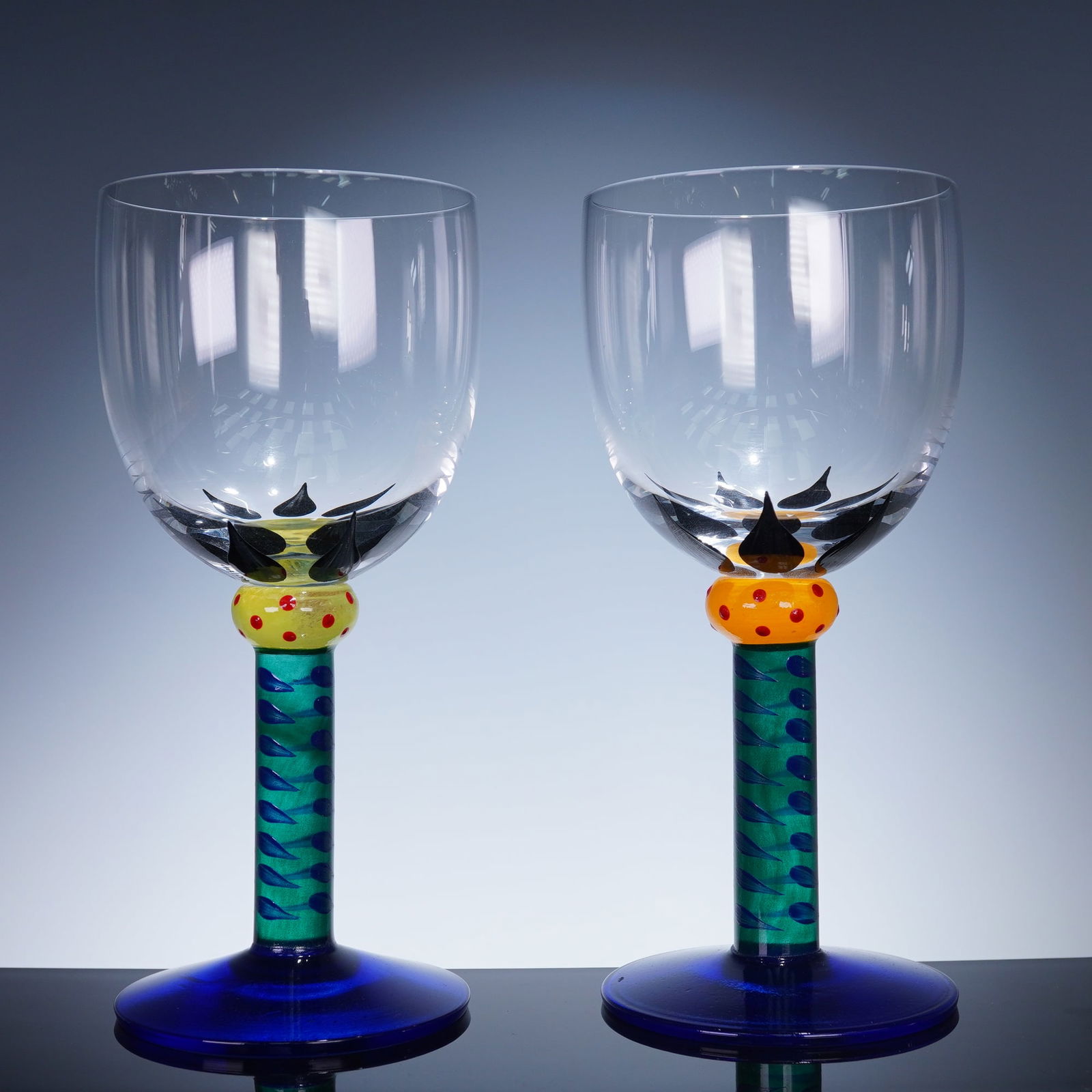 Pair of Kosta Boda Ken Done Coll Wine Glasses, Palm Trees: A pair of blown glass wine glasses hand decorated with stylized black palm tree motifs, accented at the stem junction with vividly colored knops, one in yellow with red dots and the other in orange wi