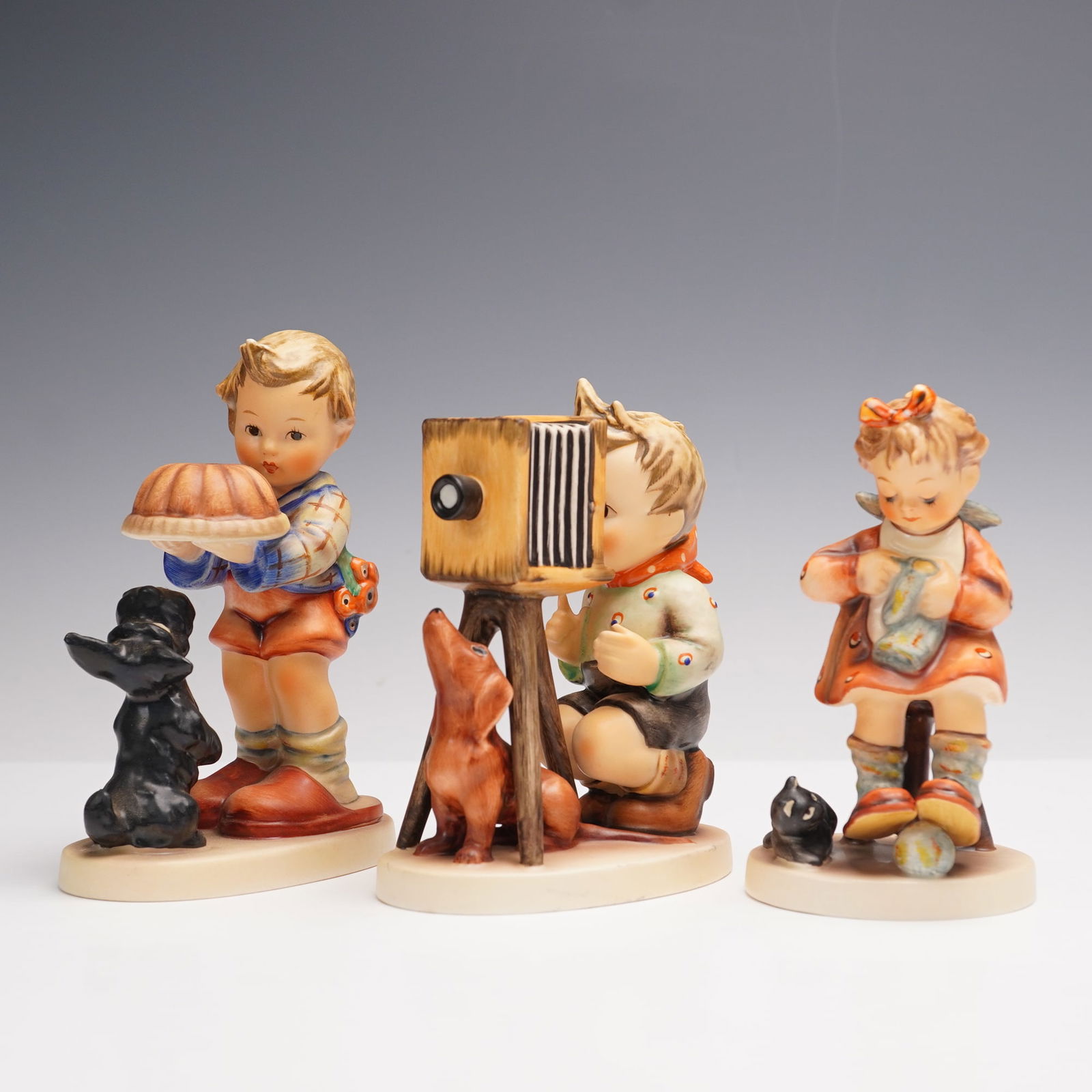 3pc Goebel Hummel Hobbyist Figurines: Hobby Figurines including The Photographer (178), depicting a young boy with an antique-style camera and dogs at his feet, Begging His Share (9), showing a boy offering a cake while a small dog looks