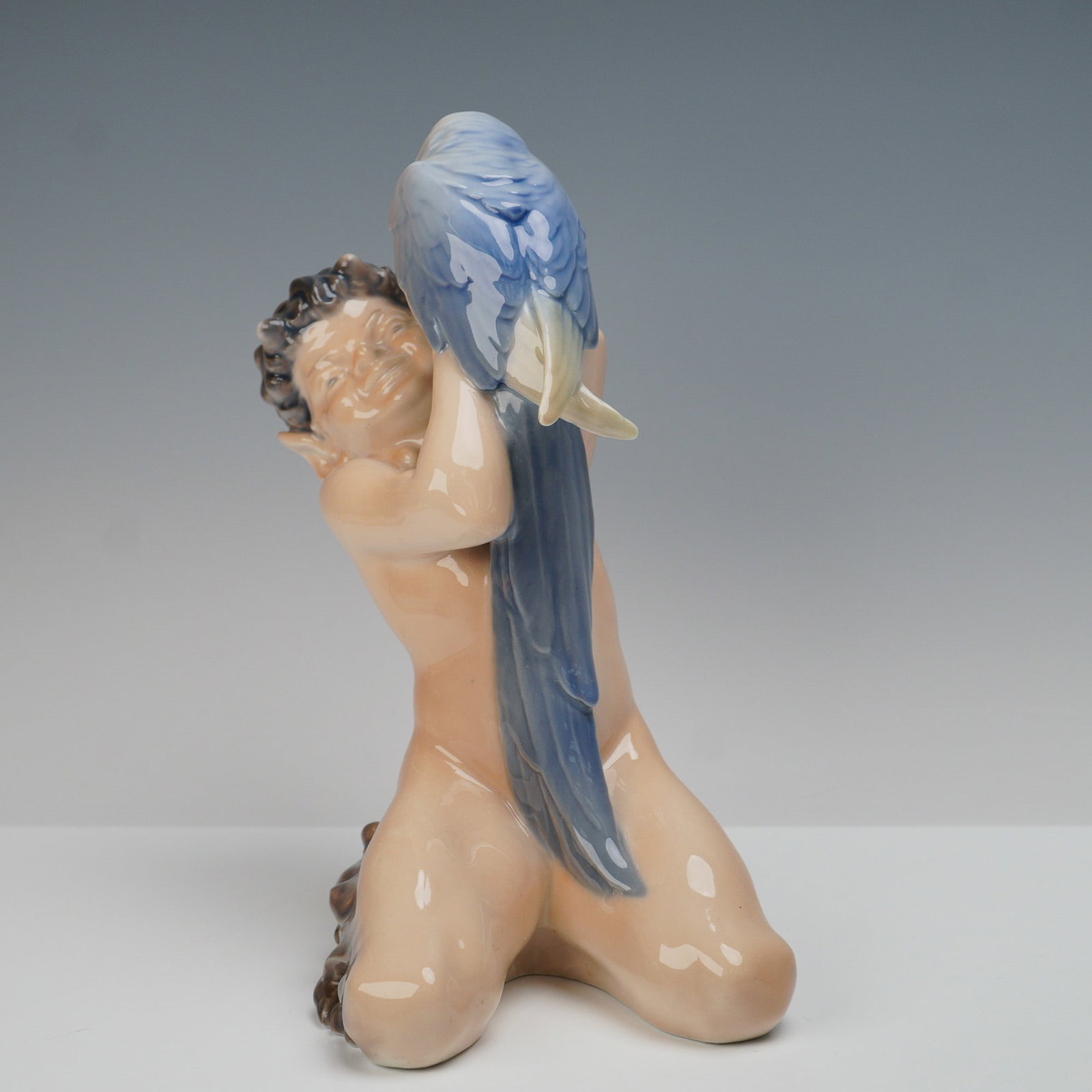 Royal Copenhagen Figurine, Faun With Parrot 752: A finely crafted porcelain figurine depicting a young faun seated on his knees while holding a large parrot perched affectionately on his arms. The faun is rendered with detailed facial features, curl