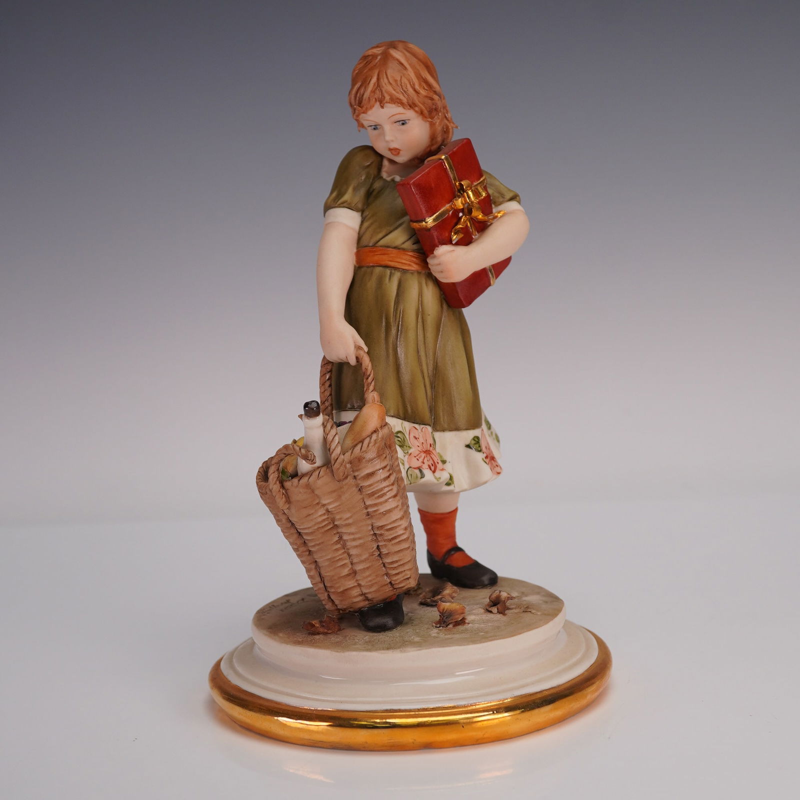 Sandro Maggioni Capodimonte Porcelain Figurine, Girl with Basket: Charming in detail, this Capodimonte porcelain figurine by Sandro Maggioni, dated 1978, depicts a young girl carrying a basket of groceries with fruit, bread, and a bottle, while holding a wrapped gif