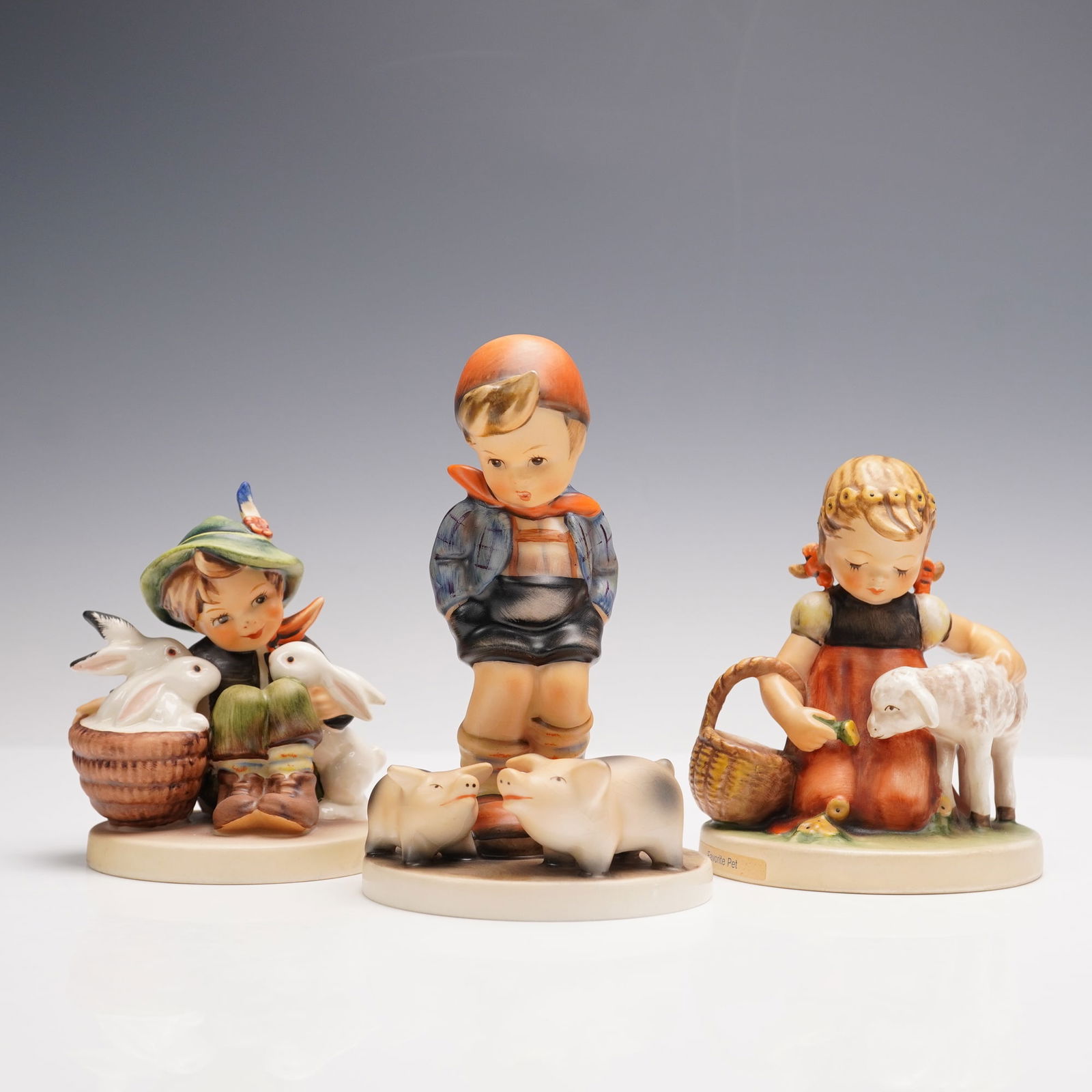 3pc Goebel Hummel Figurines, Children with Animals: A sweet trio of Goebel Hummel porcelain figurines showing children with their farmyard companions. One depicts a boy seated with a basket of rabbits, another shows a young boy standing beside two play