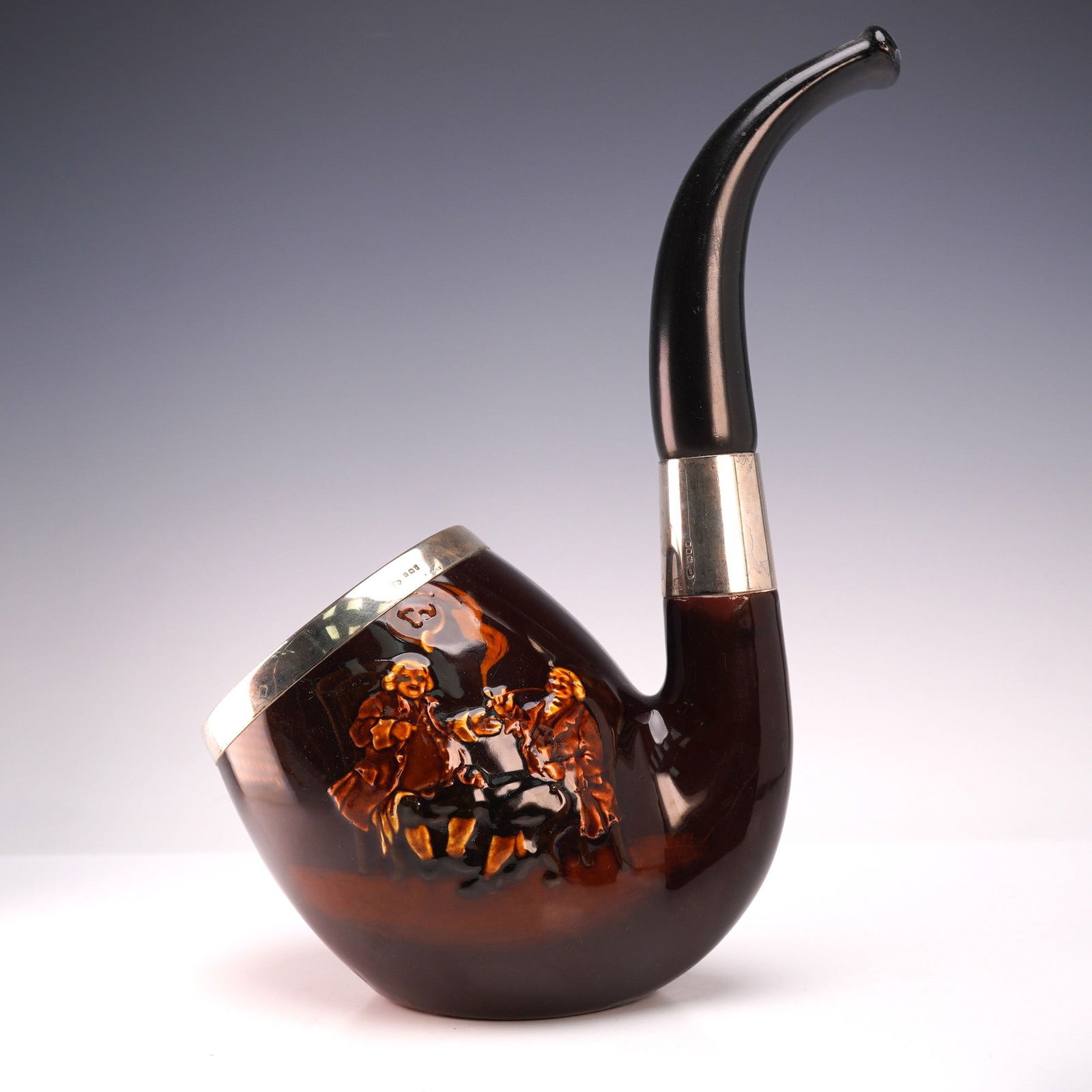 Royal Doulton Kingsware Novelty Smoking Pipe: A rare Royal Doulton Kingsware novelty smoking pipe, decorated with a raised tavern scene in rich brown glaze. The pipe is fitted with silver mounts hallmarked for Birmingham, with maker's mark visibl