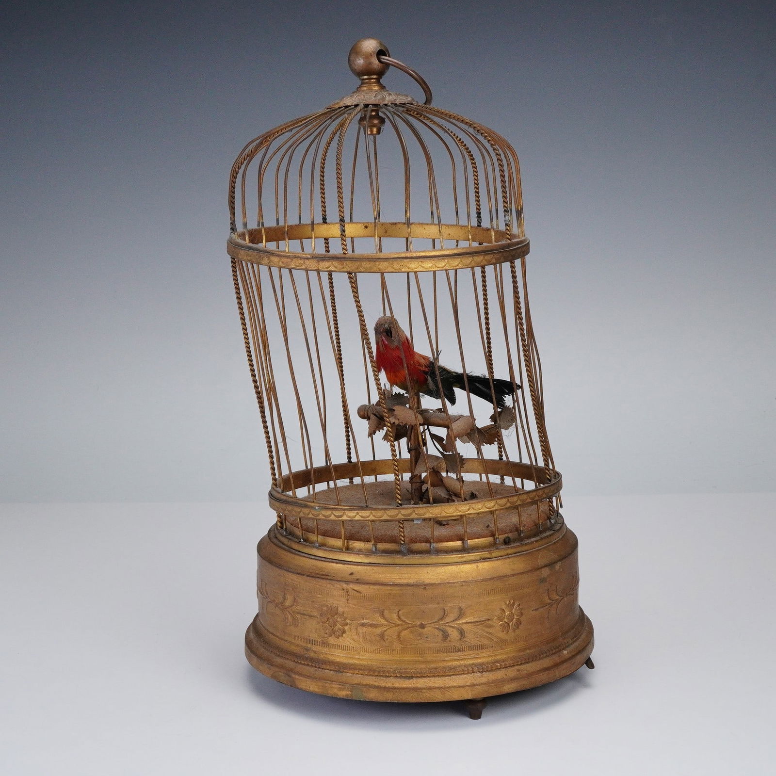 Gilt Birdcage Music Box: A charming gilt cage houses a vivid bird with bright red and black plumage perched on a leafy branch. The ornate base is richly decorated with floral patterns and conceals the music box mechanism unde