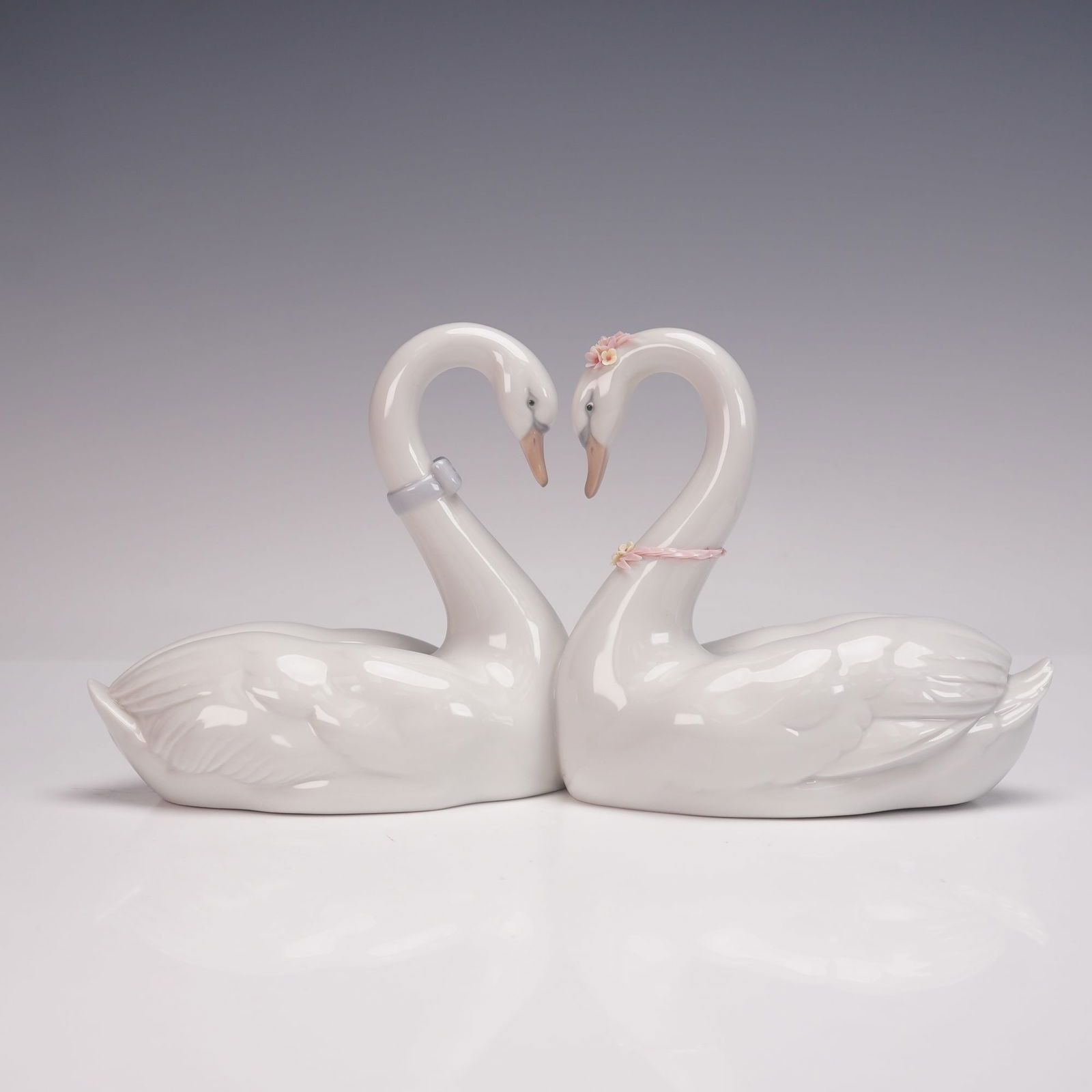Lladro Porcelain Figurine, Endless Love 1006585: Glossy finish figure modeled as swans with gracefully arched necks forming a mirrored composition. One swan is adorned with a pale blue ribbon collar, while the other bears applied pink porcelain flow