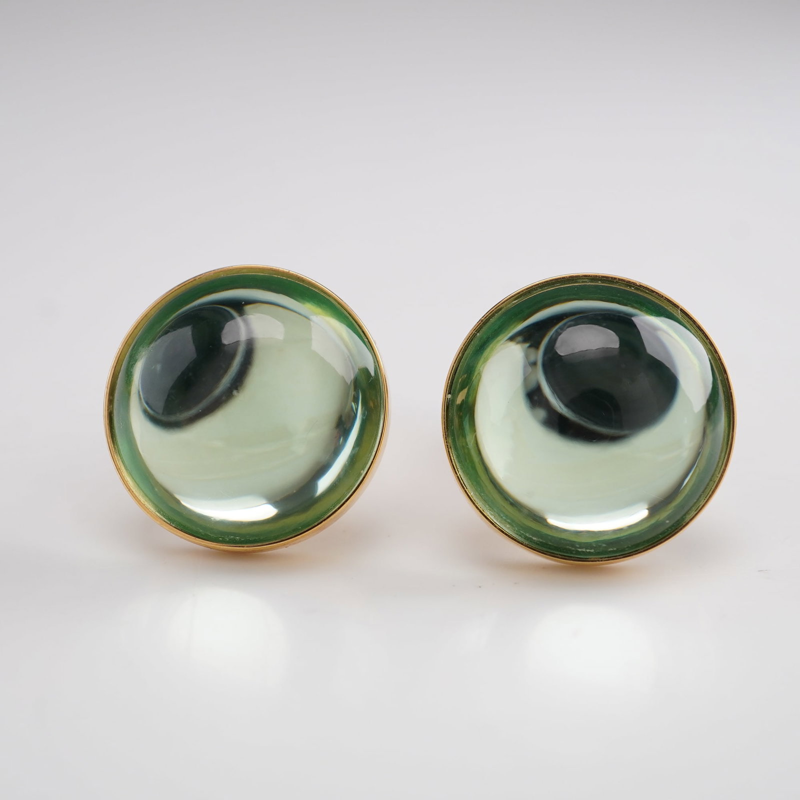 Lalique Crystal Green Cabochon Clip-On Earrings: Elegant Lalique clip-on earrings featuring round green crystal cabochons with a domed, magnifying effect, set in gold-tone mounts. The translucent crystal displays subtle green tones that shift with t