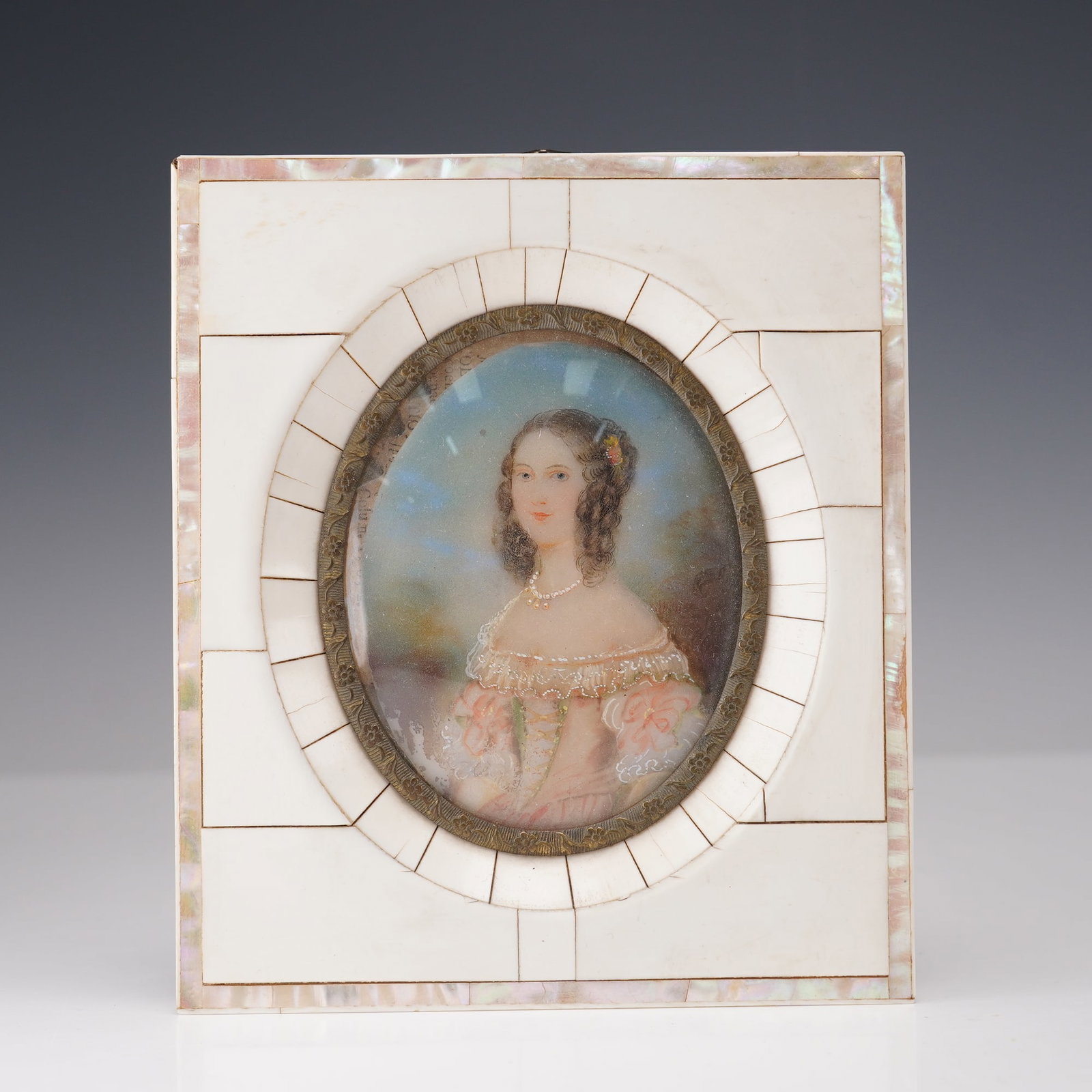 Miniature Portrait in Mother-of-Pearl and Bone Frame: A finely hand-painted 19th century miniature portrait depicting a young woman with dark curled hair adorned with a small flower, wearing an off-the-shoulder pink gown with lace trim and pearl necklace