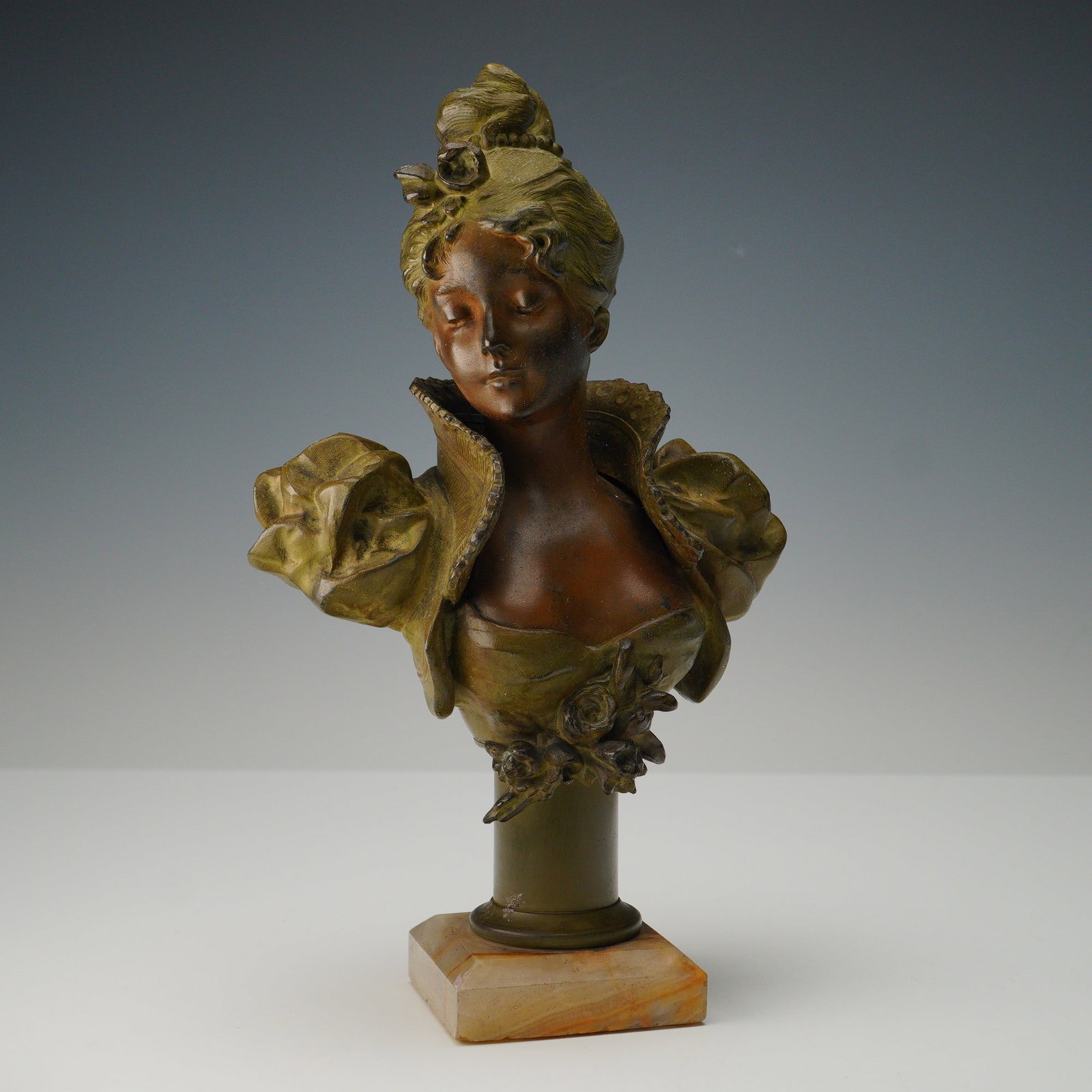 Spelter Bust of a Lady on Onyx Base: Bust depicting a refined lady with eyes gently closed, her hair arranged in an elaborate updo adorned with a rose. She wears a high-collared gown with dramatic puffed sleeves and floral embellishment 