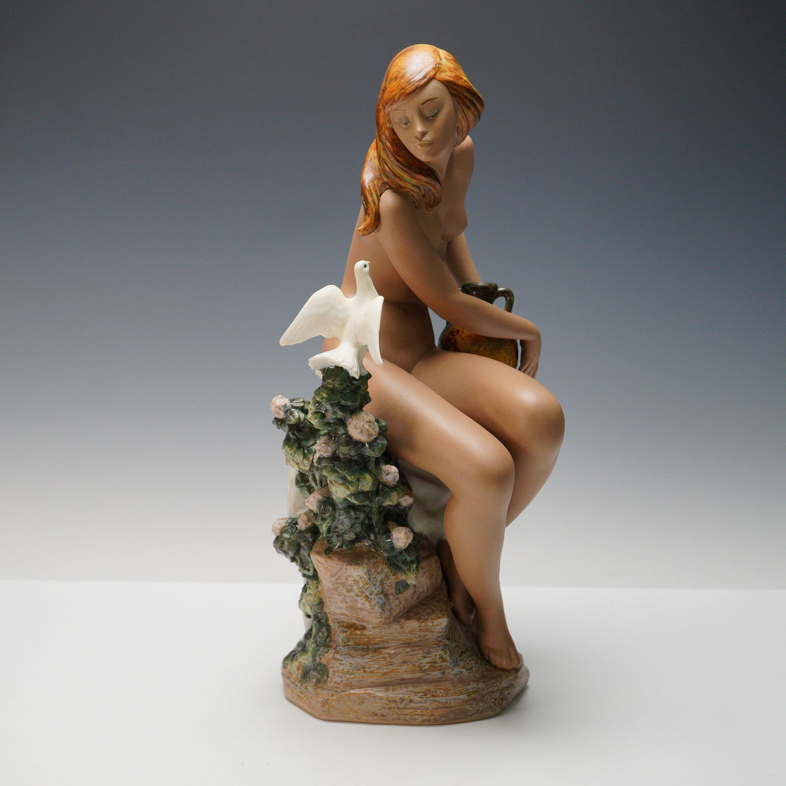 Lladro Porcelain Sculpture, Nude with Dove 1013503: A finely crafted Lladro porcelain figurine in Gres finish, depicting a nude maiden seated on a naturalistic rocky base with flowering foliage, a dove perched before her and a rustic water jug resting