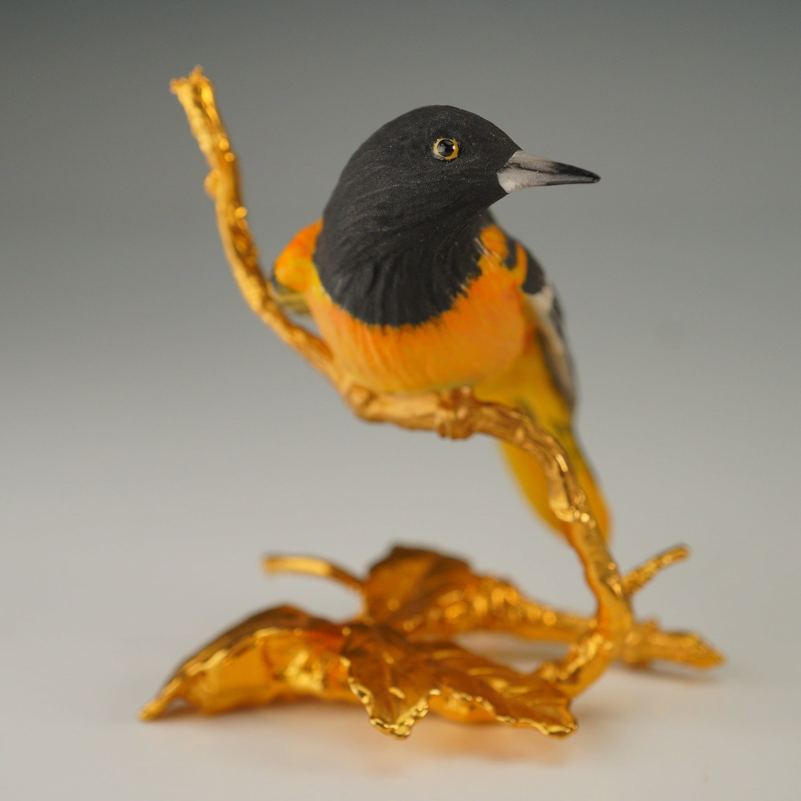 Boehm Miniature Porcelain Figurine, Oriole Perched on Gold Base: A finely detailed porcelain figurine depicting an Oriole bird on a gilt branch. The bird is rendered in striking tones of black and vivid orange with sharply delineated wing markings, and finished wit