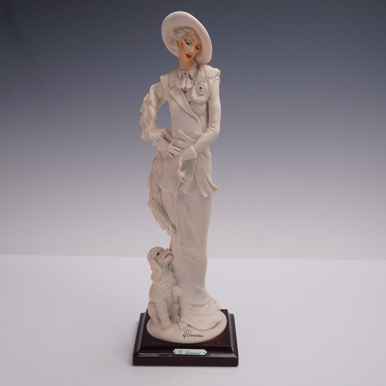 Florence Giuseppe Armani Figurine, Lady with Dog: A figurine depicting an elegantly dressed lady wearing a wide-brimmed hat and long coat, with a poodle at her side. Signed G. Armani on the base with the Florence stamp. Mounted on a square wooden bas