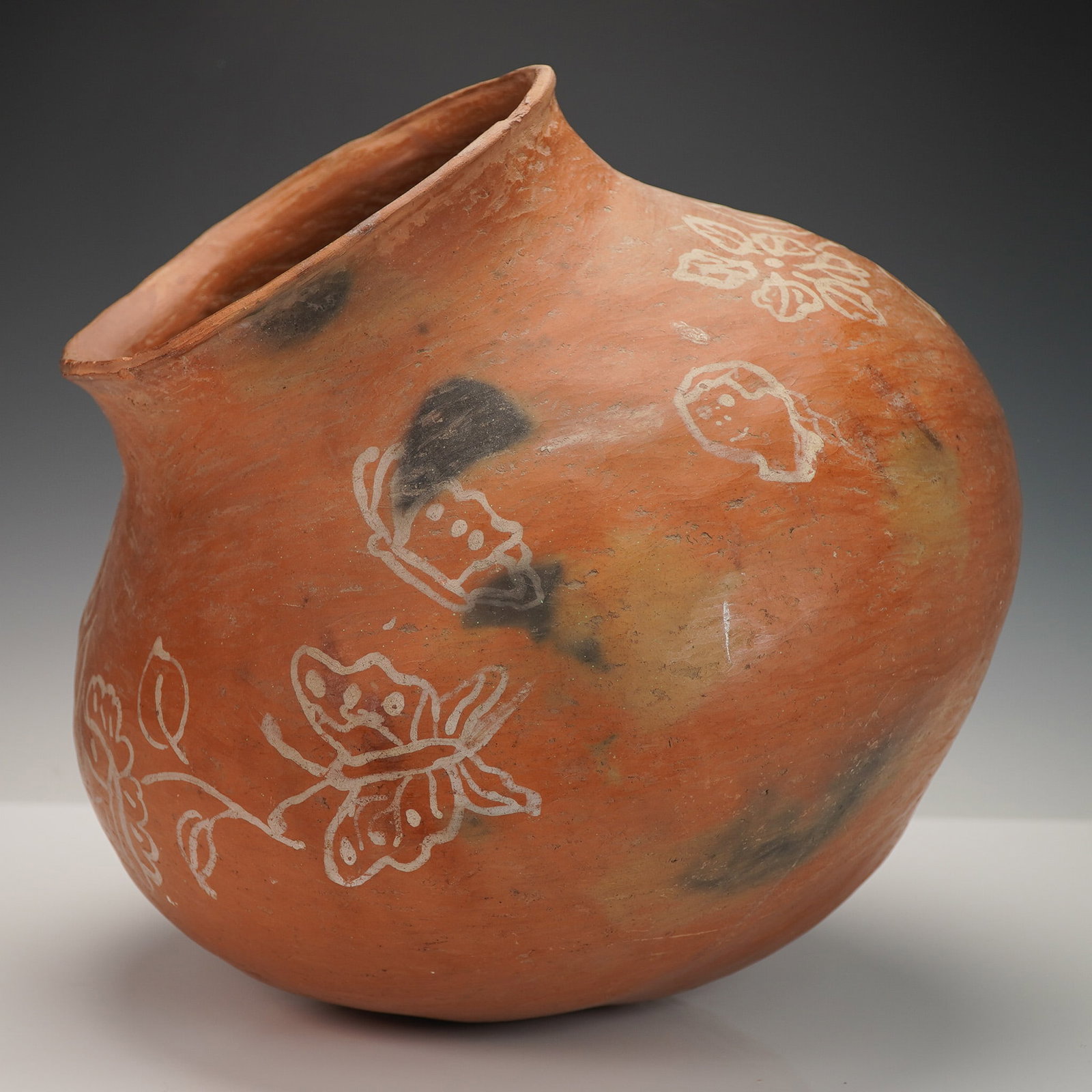 Mesoamerican Red Clay Water Pot with Butterflies and Floral Designs: This red clay water pot is decorated with white painted butterflies and floral motifs. The pot shows visible signs of age and use, including multiple cracks and surface wear, particularly along the bo