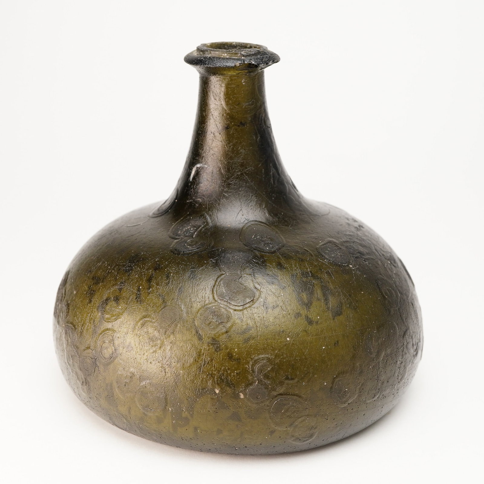 Antique Green Glass Onion Wine Bottle: An onion wine bottle in olive-green glass, featuring a characteristically squat, bulbous form with a short neck and wide base. The surface displays both carved and embossed circular splotch patterns d