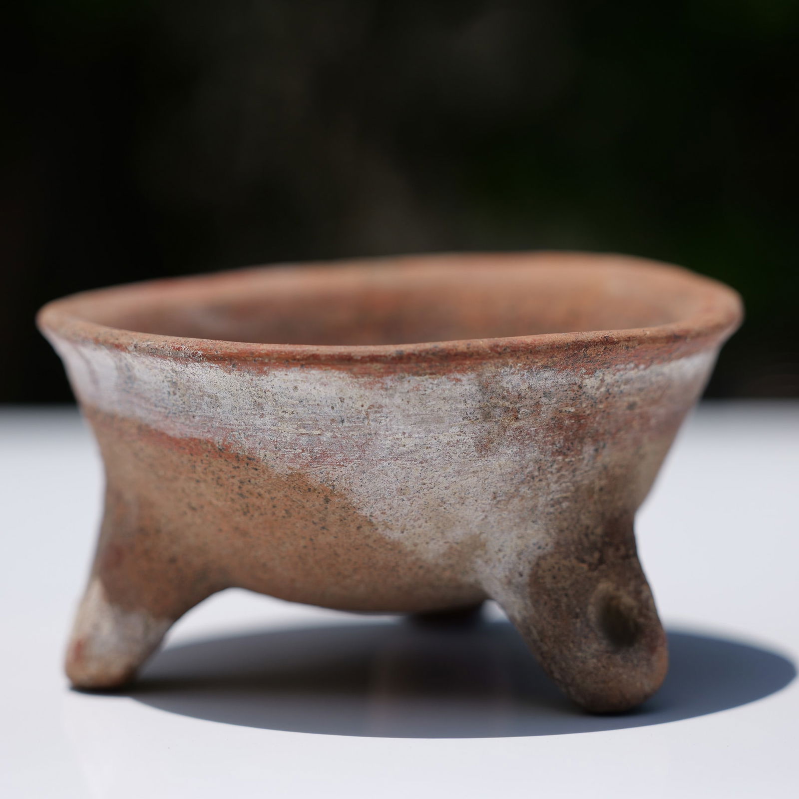 Pre-Columbian Redware Tripod Bowl: It is constructed from reddish-brown earthenware and supported by three stubby legs that terminate in pierced lugs, which may have served a suspension or binding function. The legs are integrated into