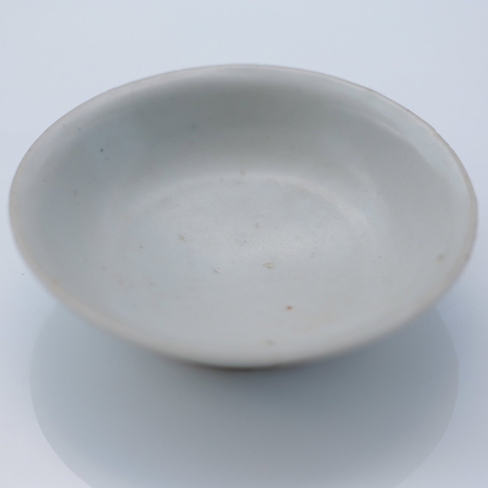 Korean White Glazed Porcelain Saucer: A porcelain saucer with a white glaze finish. The saucer exhibits a smooth, rounded form with slightly upturned edges and a shallow central basin. The glaze is thin and subtly translucent in areas, re