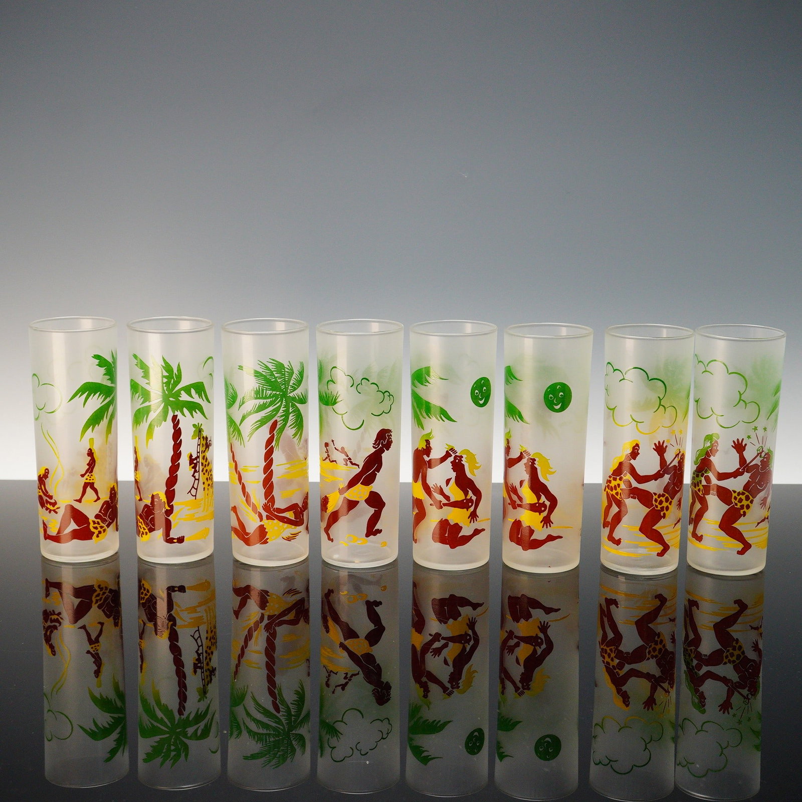 8pc Federal Glass Frosted Highball Glasses with Cavepeople Motif: A colorful mid-century set of 8 frosted highball glasses by Federal Glass, each decorated with playful cavewoman and caveman scenes among palm trees and clouds. The bold red, green, and yellow enamel 
