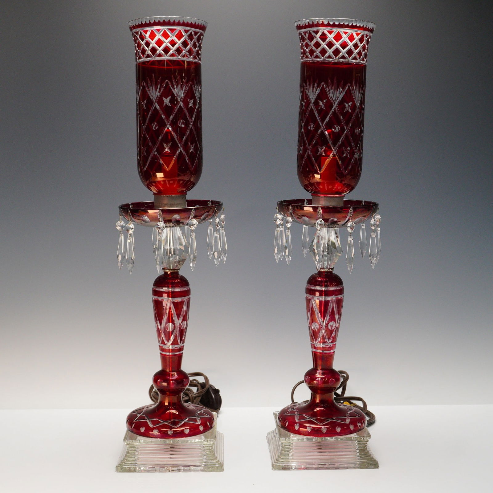 Pair of Bohemian Cut-to-Clear Red Glass Mantle Lusters with Crystal Prisms: This stunning pair of Bohemian mantle lusters features elegant cut-to-clear red glass with intricate diamond and star patterns. The lamps are set on square glass bases and adorned with sparkling cryst