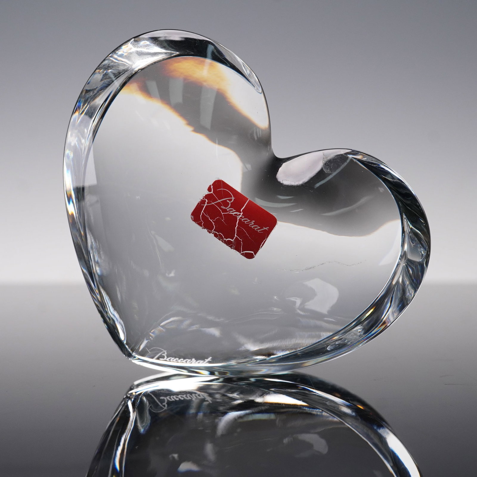 Baccarat Crystal Heart Paperweight: Clear Baccarat crystal heart shaped paperweight with red Baccarat label intact. Elegant design with smooth faceted edges, signed Baccarat on base. Measures 3.5"L x 1.75"H. Issued: 20th century Dimensi