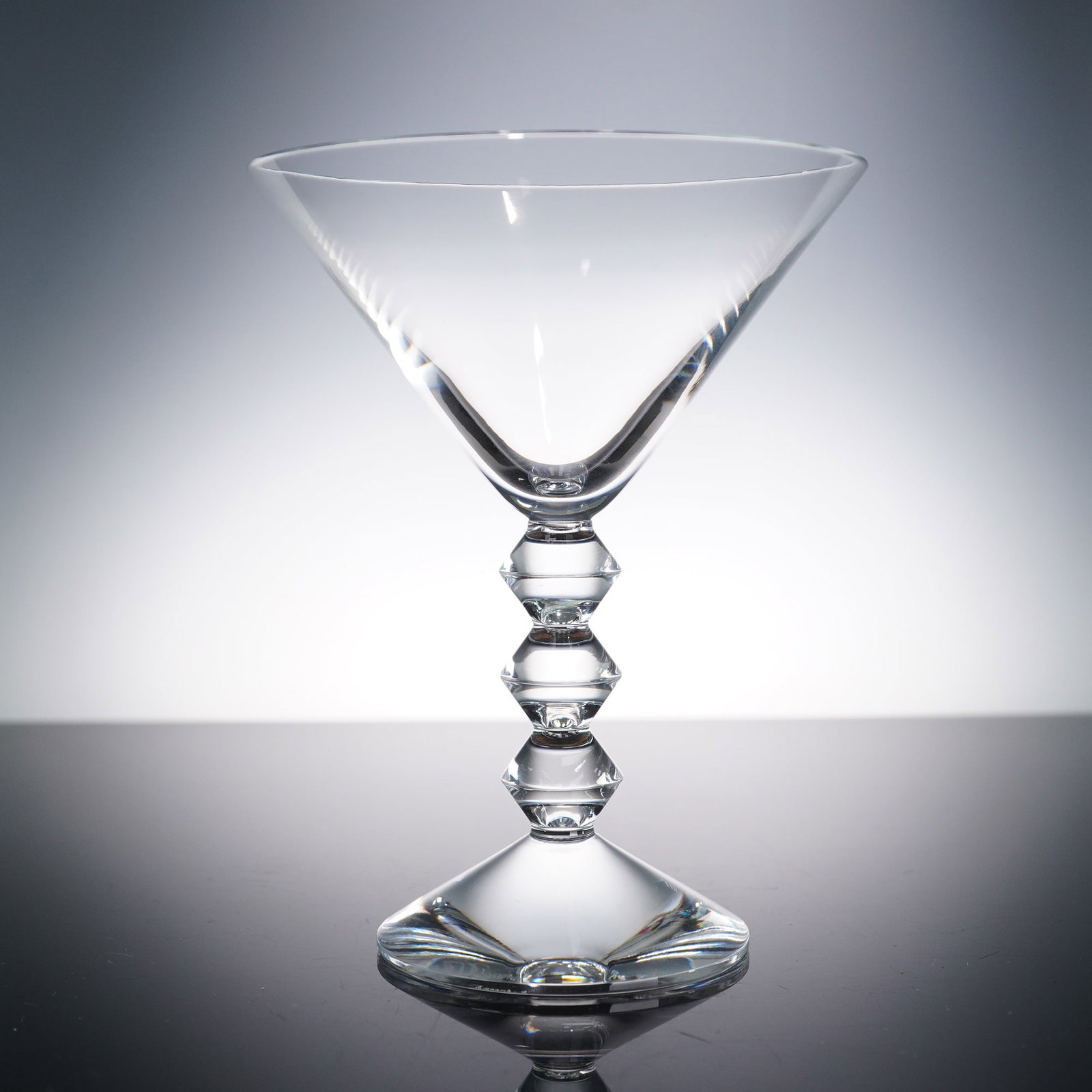 Baccarat Vega Martini Glass: Martini glass, featuring a striking contemporary design with a clear, faceted stem composed of stacked diamond-shaped elements and a broad, conical bowl. Crafted in fine crystal, this elegant glass sh
