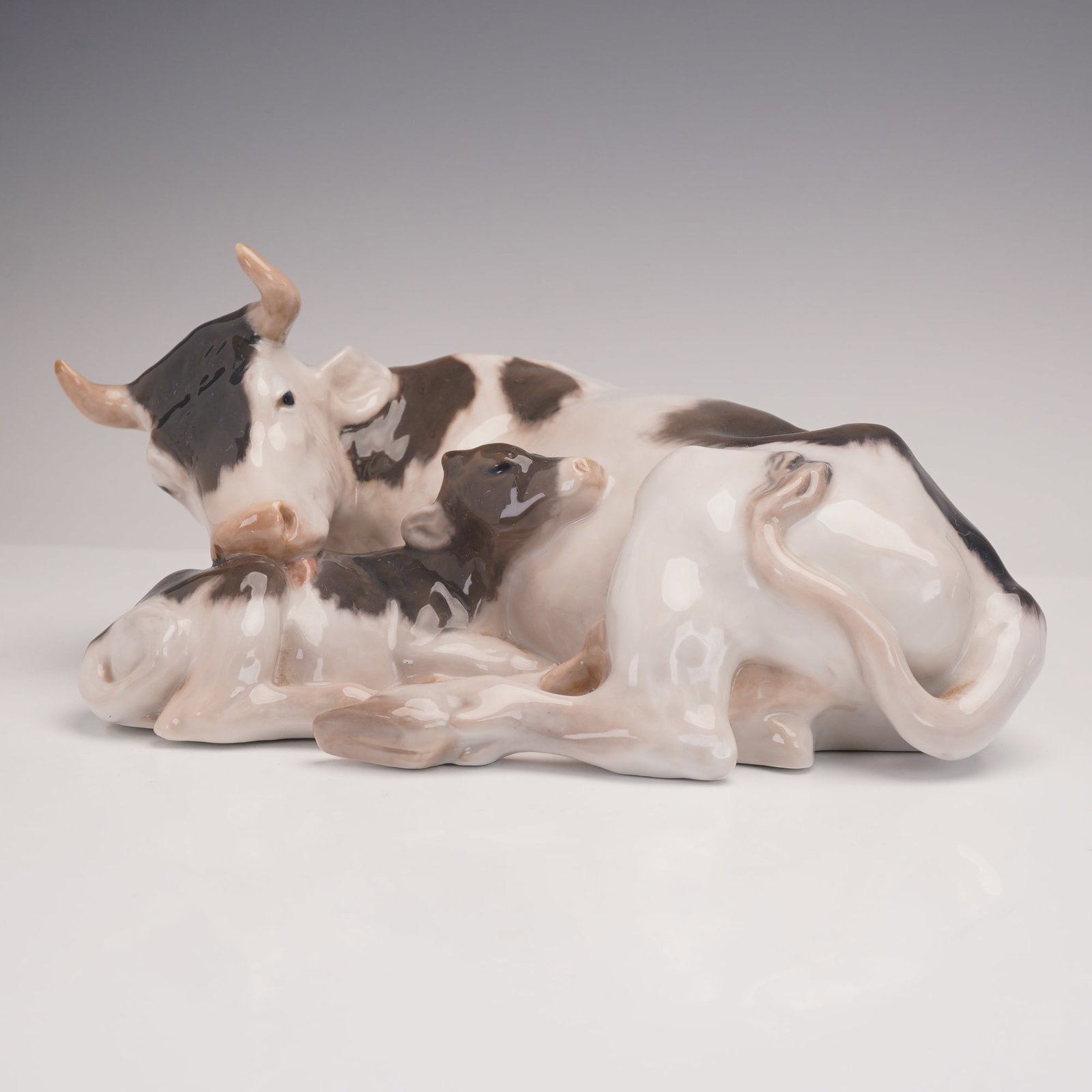Royal Copenhagen Porcelain Figurine, Cow with Calf 800: A touching Royal Copenhagen porcelain group depicting a cow tenderly nuzzling her calf as they rest together. The naturalistic glaze in shades of brown, black, and cream captures the lifelike markings
