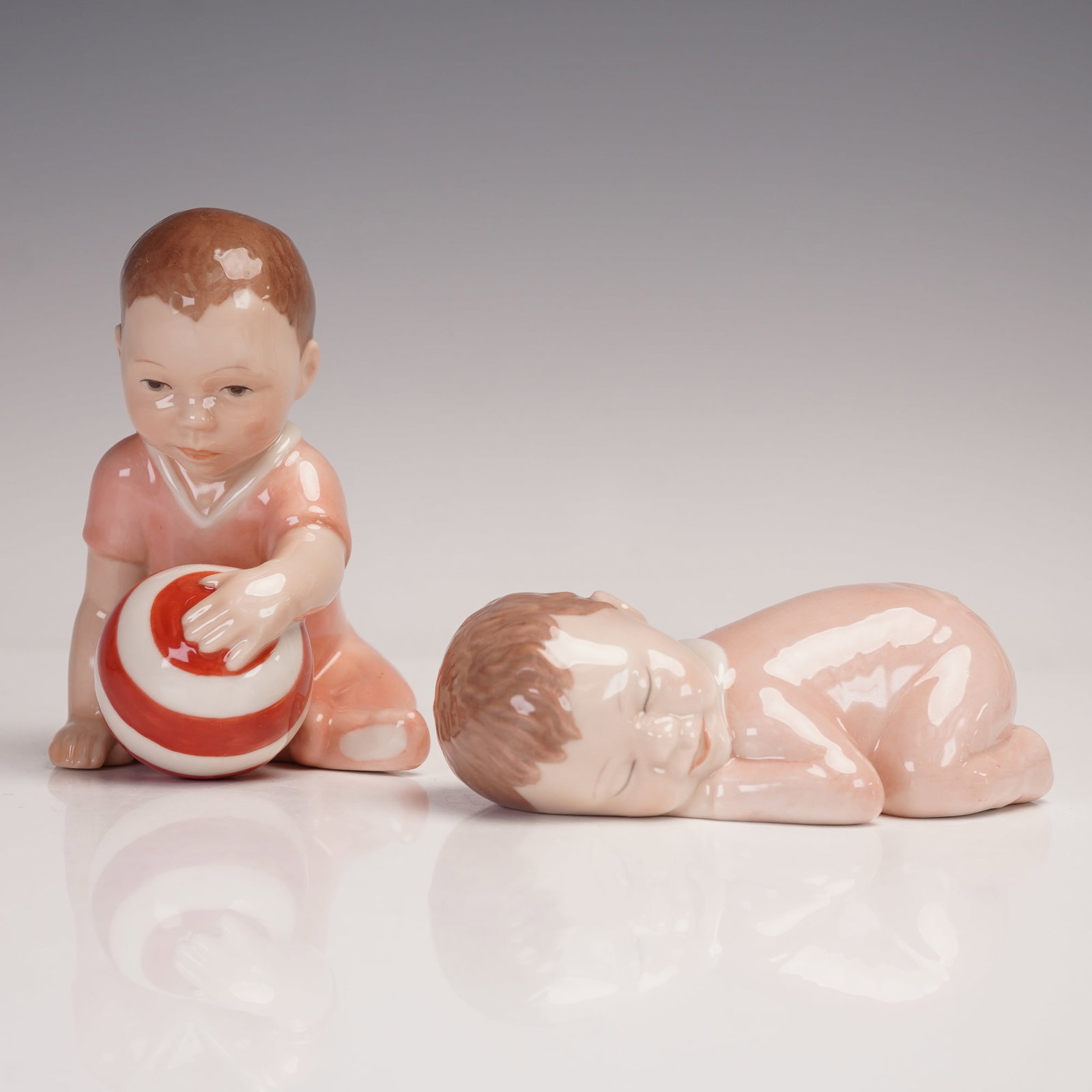 2pc Royal Copenhagen Porcelain Figurines, Boy With Ball & Sleeping Infant: A charming pair of Royal Copenhagen porcelain figurines featuring tender childhood scenes. The first depicts a young boy in soft peach attire playing with a bold red and white striped ball, model 023,