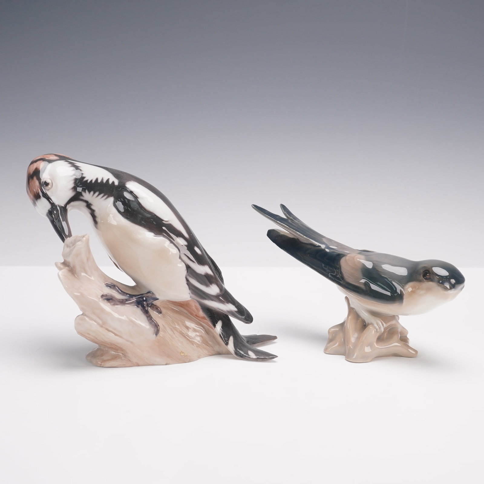 2pc Bing & Grondahl Porcelain Bird Figurines, Woodpecker & Swallow: A pair of hand-painted porcelain bird figurines by Bing & Grondahl, Denmark. The group includes a finely modeled woodpecker perched on a tree stump, rendered with naturalistic black, white, and russet