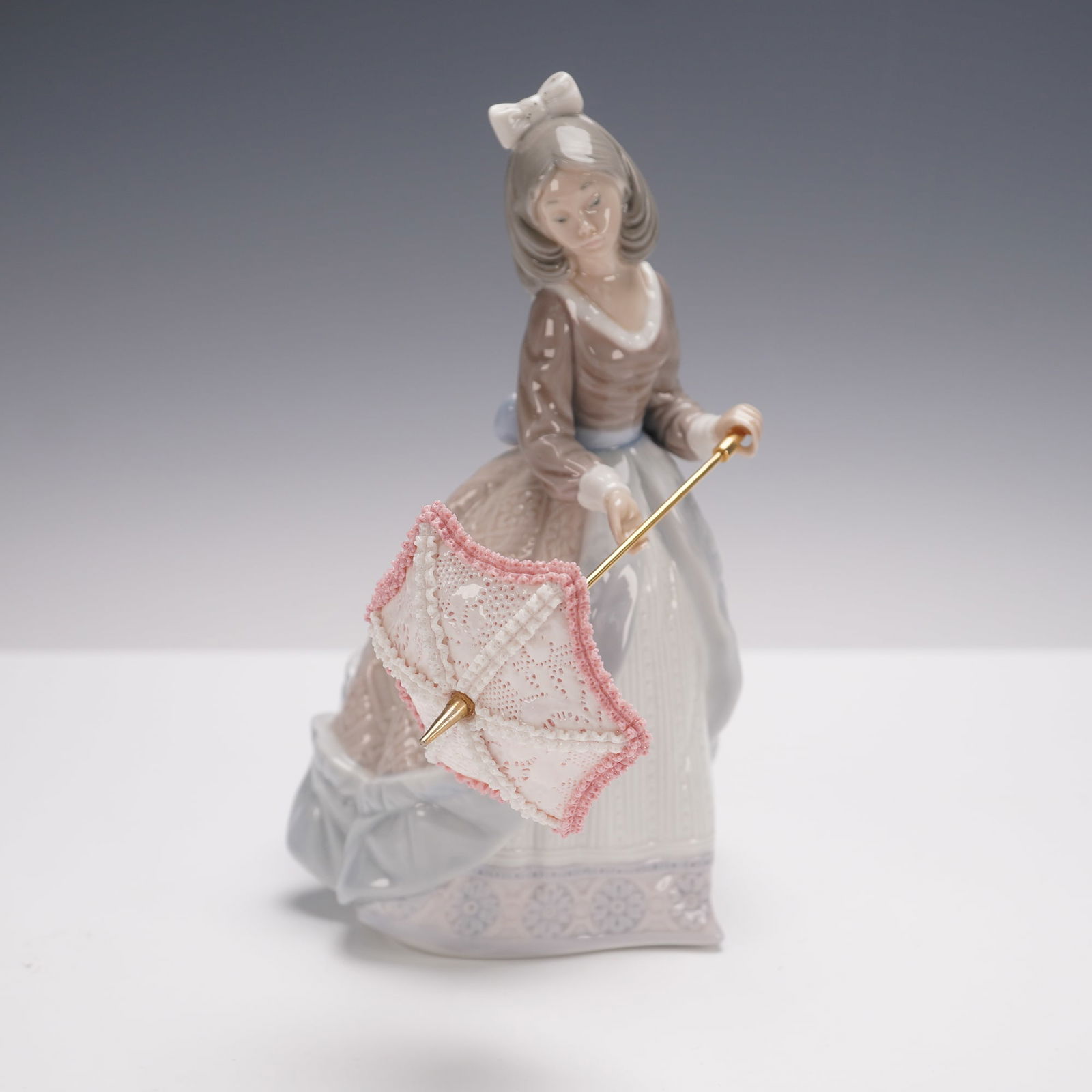 Lladro Porcelain Figurine, Jolie 1005210: Glossy finish figure depicting a young girl in a flowing gown holding an intricately formed parasol. The figure is adorned with delicate detailing, including a textured patterned skirt. Lladro backsta