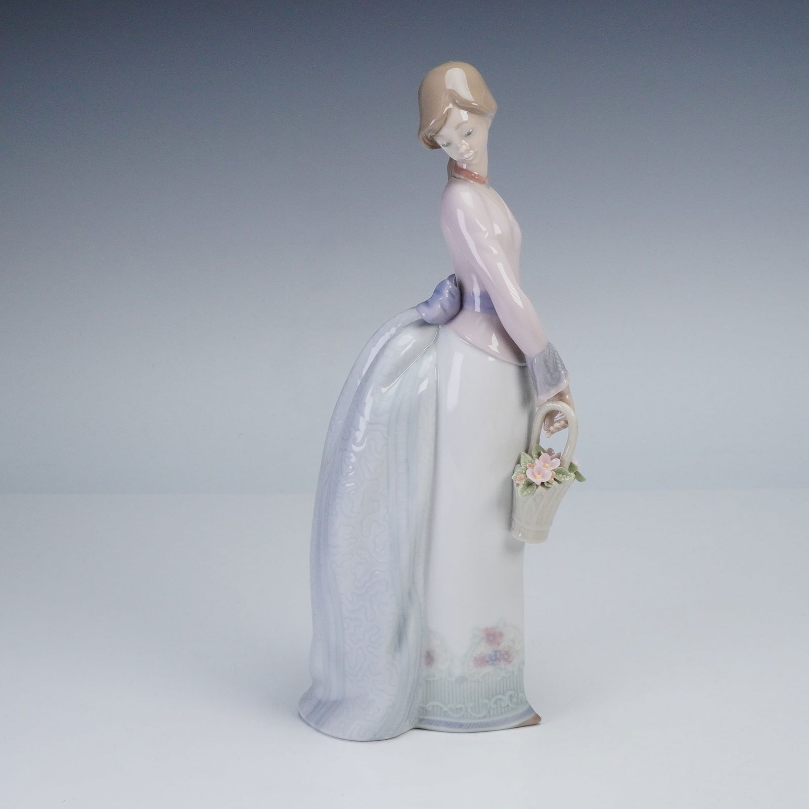 Lladro Porcelain Figurine Basket of Love 01007622: Lladro porcelain figurine titled Basket of Love, model number 01007622, depicts a graceful young woman in pastel tones carrying a basket of delicate flowers. The figure is hand-painted in soft lavende