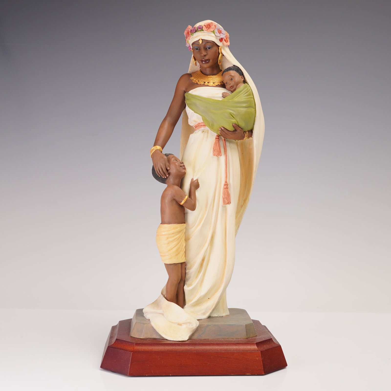 Willits Design's Ebony Vision's Figurine, The Madonna 37001: Issued as part of the Ebony Visions collection, this piece portrays a graceful woman wearing a flowing gown and floral crown, holding an infant wrapped in green cloth while a small child stands beside