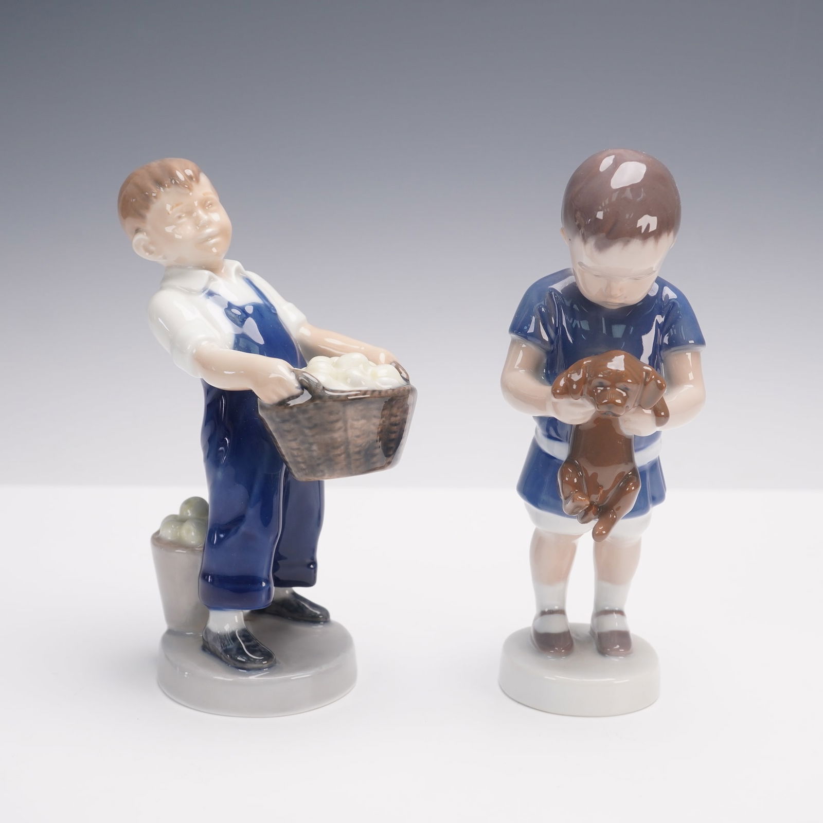 2pc Royal Copenhagen and Bing & Grondahl Porcelain Figurines: A pair of porcelain figurines including a Royal Copenhagen model of a boy carrying a basket of fruit with an additional pail beside him, and a Bing & Grondahl model of a young child holding a puppy. B