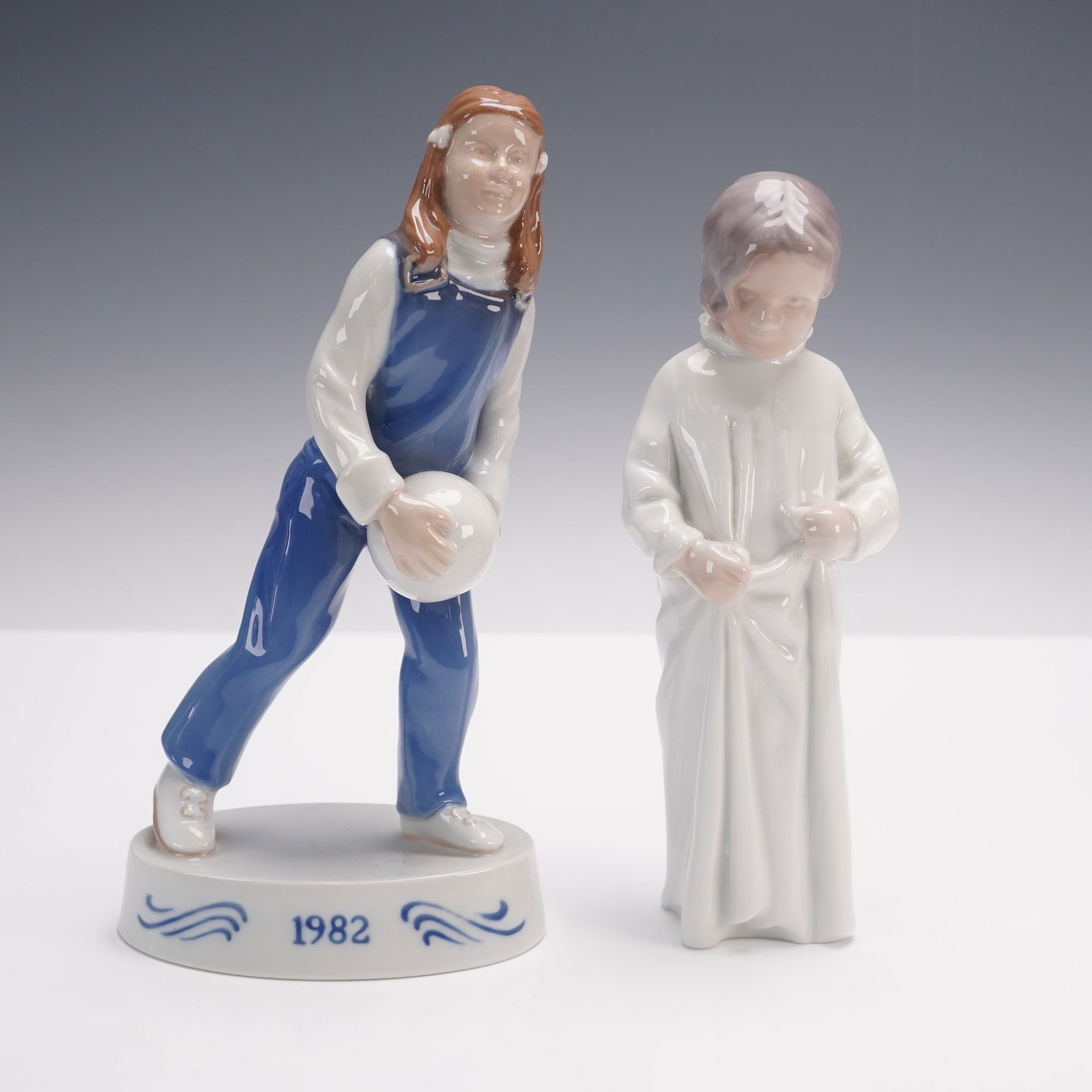 2pc Bing & Grondahl Figurines, Girl With Ball and Good Morning: One of these porcelain figurines is the 1982 Figurine of the Year titled "Girl With Ball,", depicting a young girl in blue overalls holding a ball, set on an oval base inscribed with the year. The oth