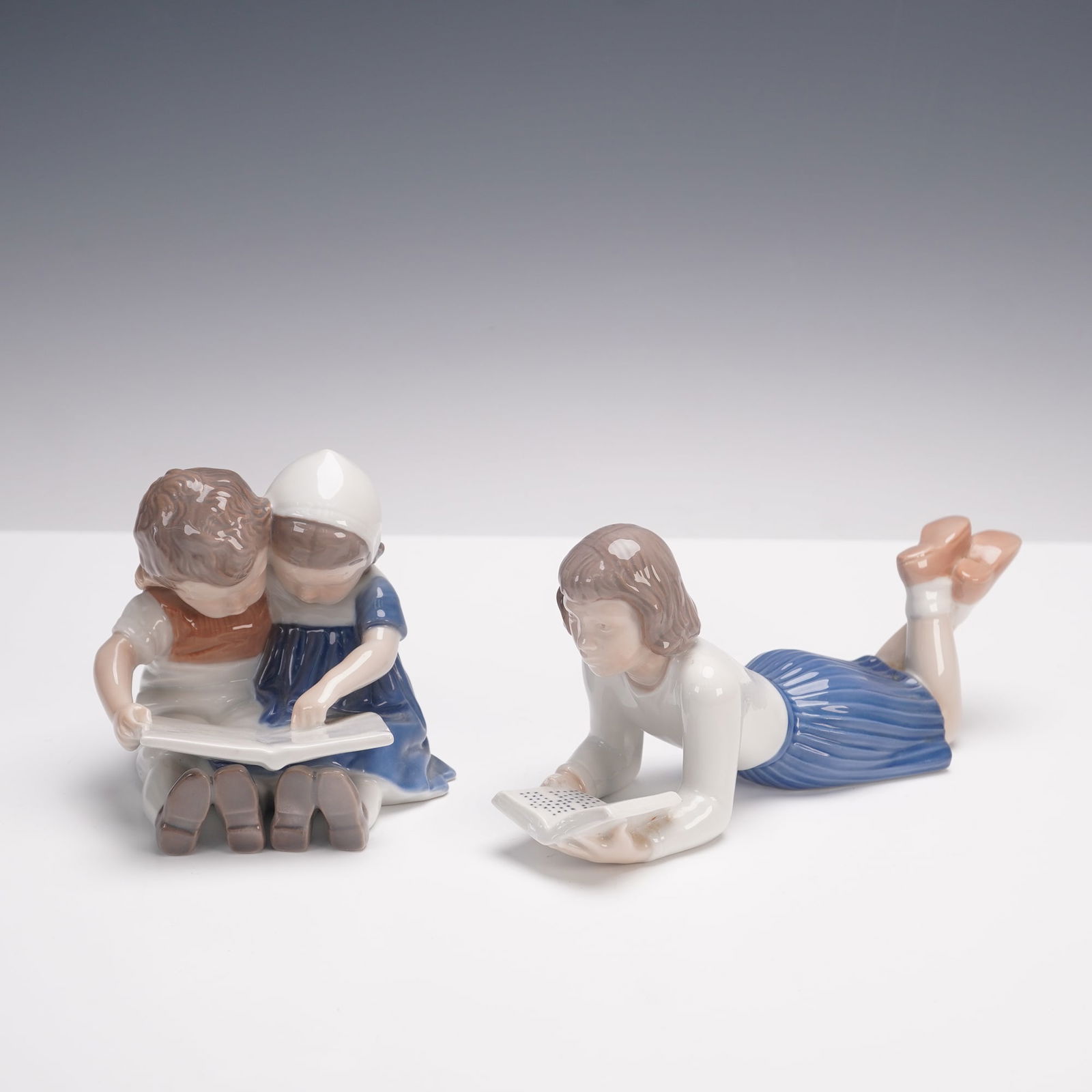 2pc Bing & Grondahl Porcelain Figurines of Children Reading: Two Bing & Grondahl porcelain figurines depicting children at study. One shows a young girl lying on her stomach reading a book, while the other portrays two children seated closely together, sharing