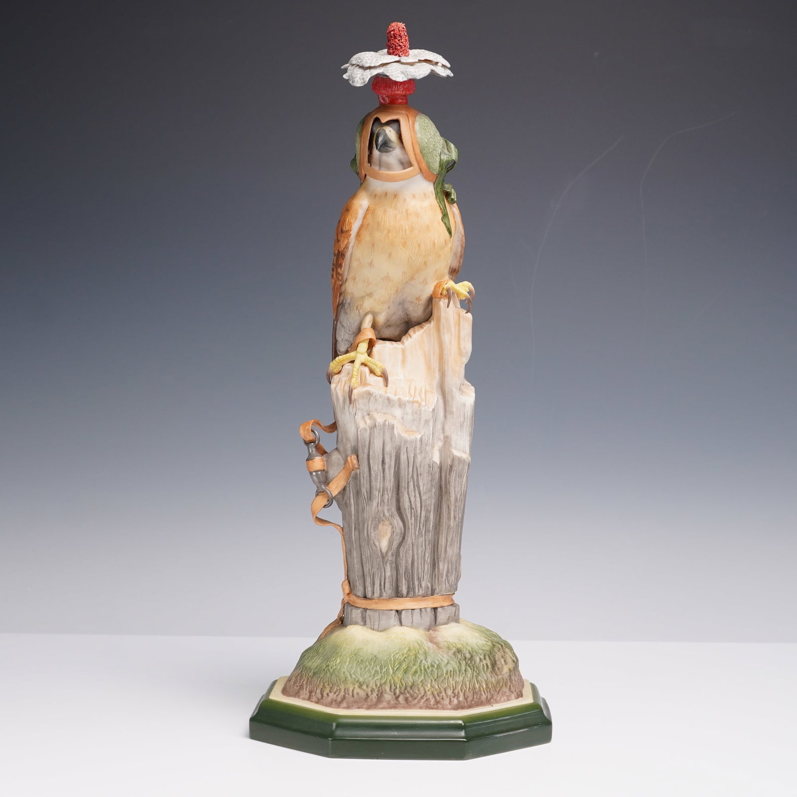 Boehm Porcelain Figurine, Kestrel 492: Depicting a kestrel perched on a detailed tree stump with leather straps and metal hardware accents, wearing a falconry hood topped with a red and white plume. The bird is hand-painted in naturalistic