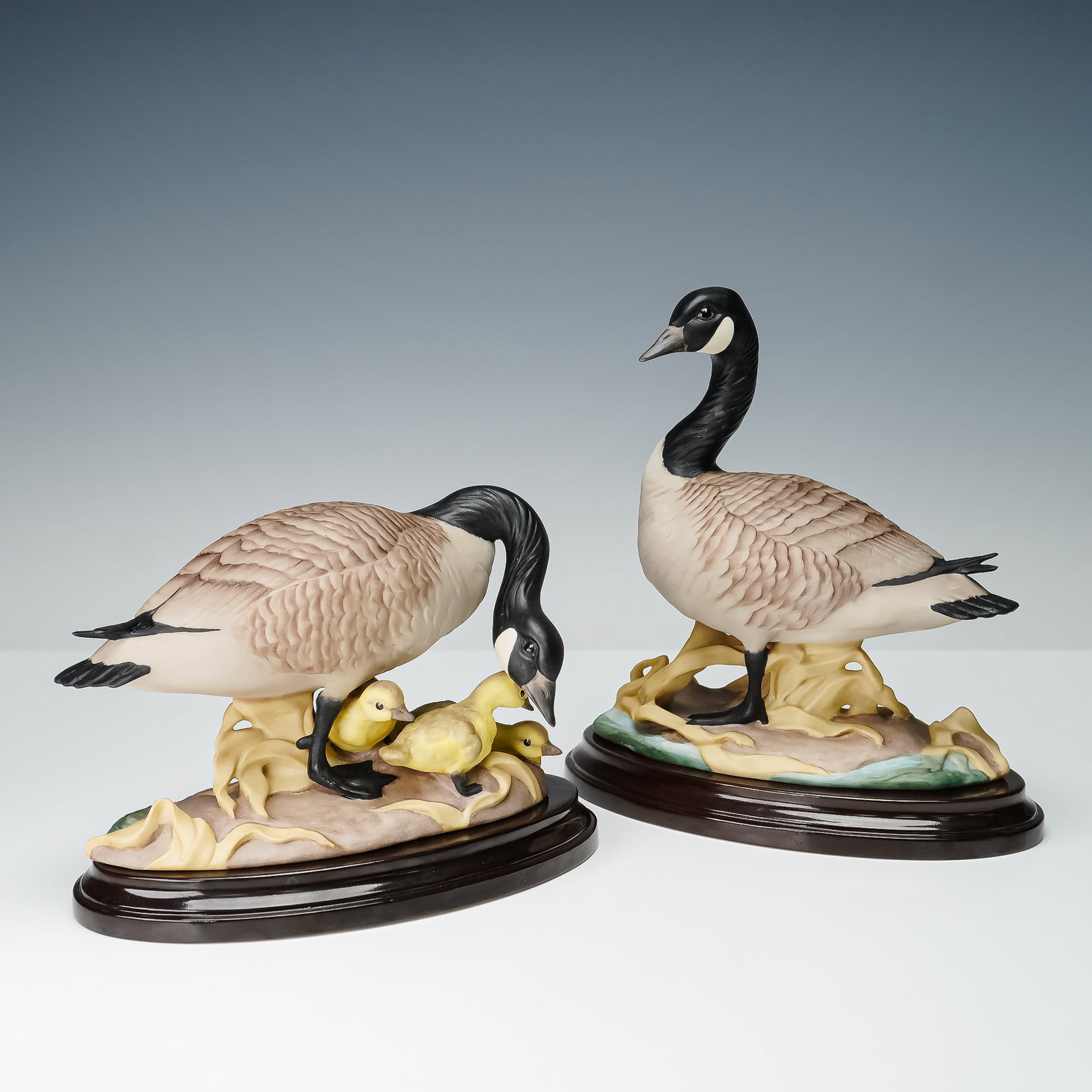 2pc Boehm Porcelain Canada Geese with Bases, 408: Pair of Boehm porcelain figurines depicting Canada geese with goslings, modeled in lifelike detail. One figure portrays a parent goose attentively positioned among its young, perched on naturalistic b