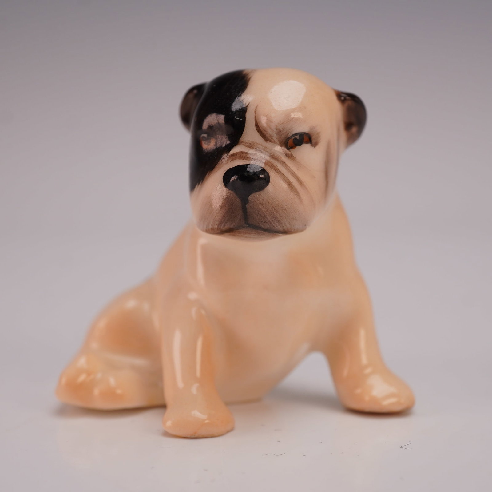 Royal Doulton Dog Figurine, Puppy Bulldog K2: A charming Royal Doulton porcelain figurine depicting a seated bulldog puppy with a tan body and distinctive black eye patch, model K2. Finely detailed and hand-painted, this piece captures the breed'