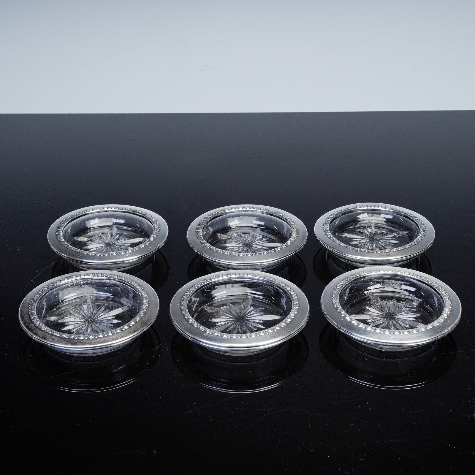6pc Sterling Silver Glass Coasters: A refined set of six round glass coasters each featuring a cut starburst design to the center. The rims are encircled with sterling silver and decorated with a repeating scalloped pattern. Gross Weigh