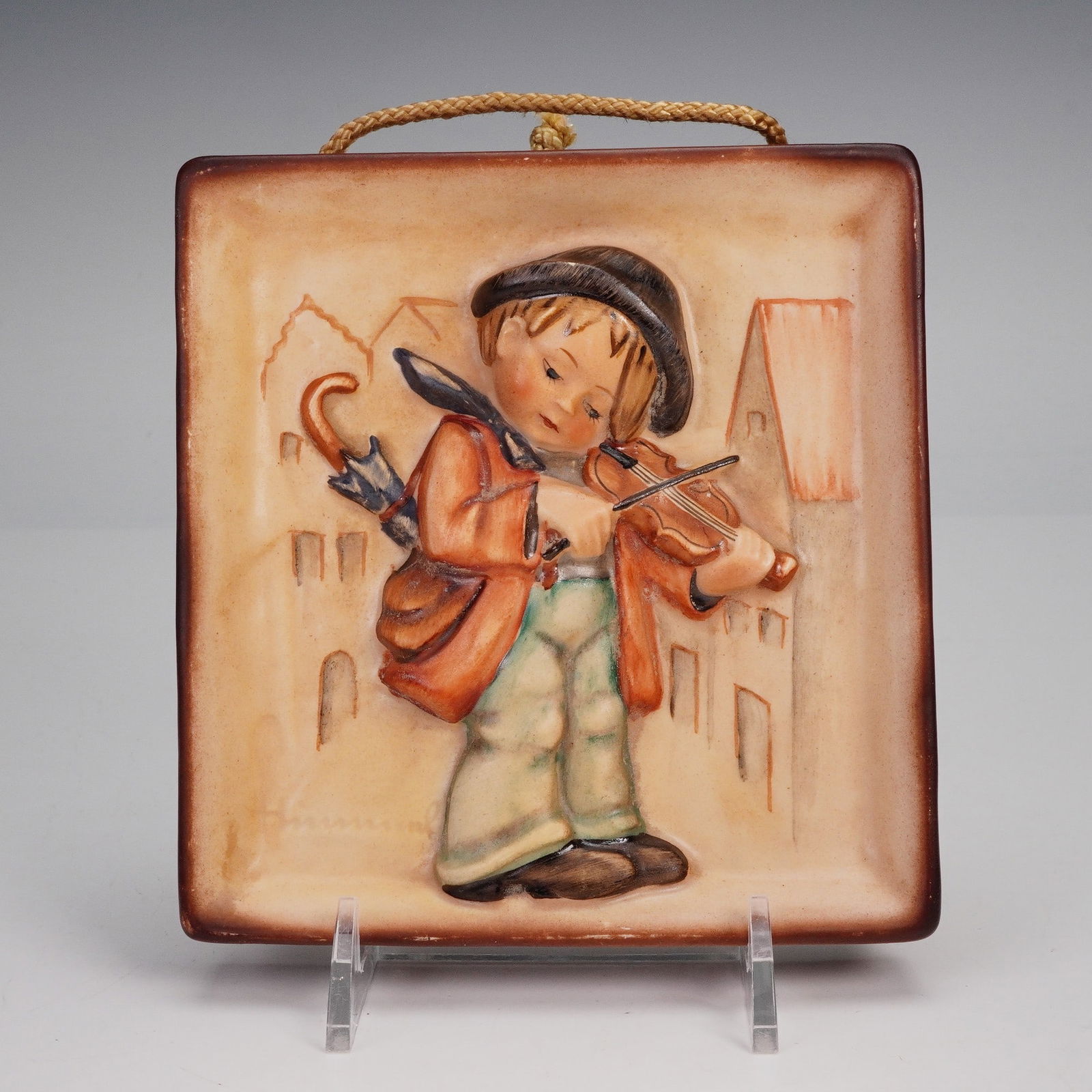 Goebel Hummel Wall Plaque, Little Fiddler: A vintage Goebel Hummel ceramic wall plaque titled Little Fiddler, featuring a young boy in traditional attire playing the violin with an umbrella tucked under his arm, set against a soft architectura