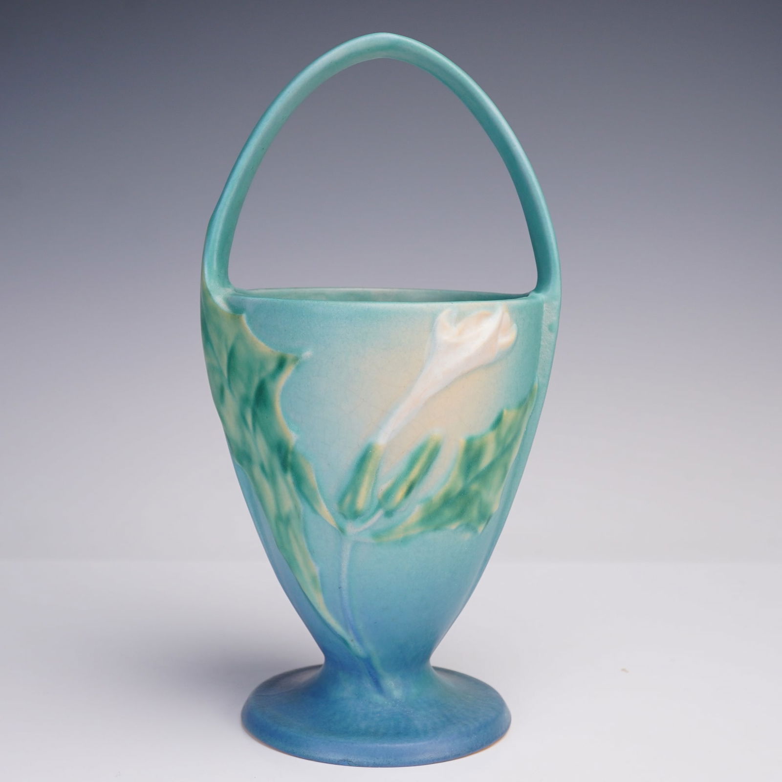 Roseville Pottery Foxglove Basket: A hand-crafted ceramic basket in the Foxglove pattern, featuring a tall arched handle and pedestal base. The exterior is decorated with raised white foxglove blossoms and green foliage set against sof